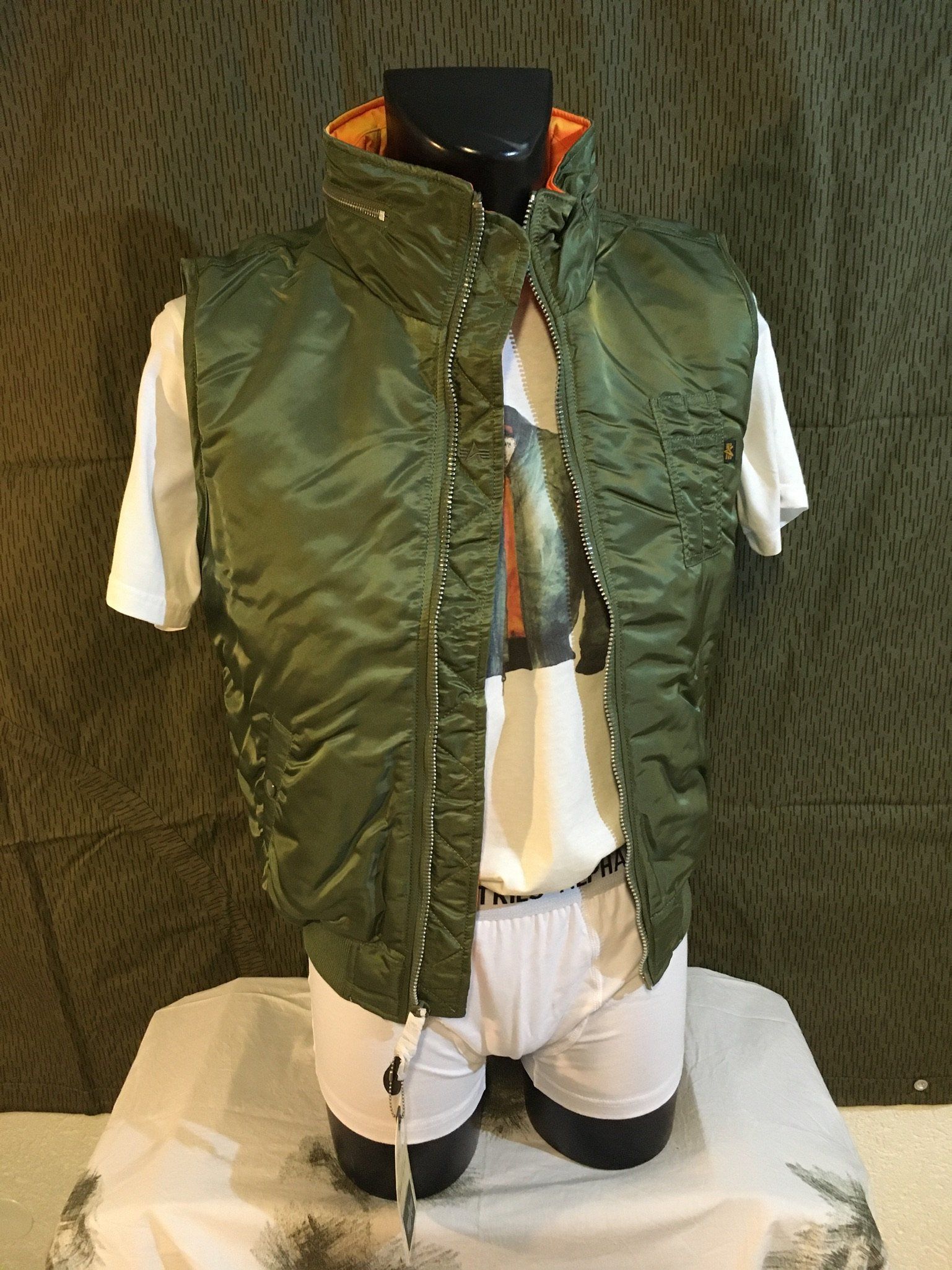 Grüne Steppweste über weißem T-Shirt auf einer schwarzen Schaufensterpuppe, mit orangefarbenem Futter.