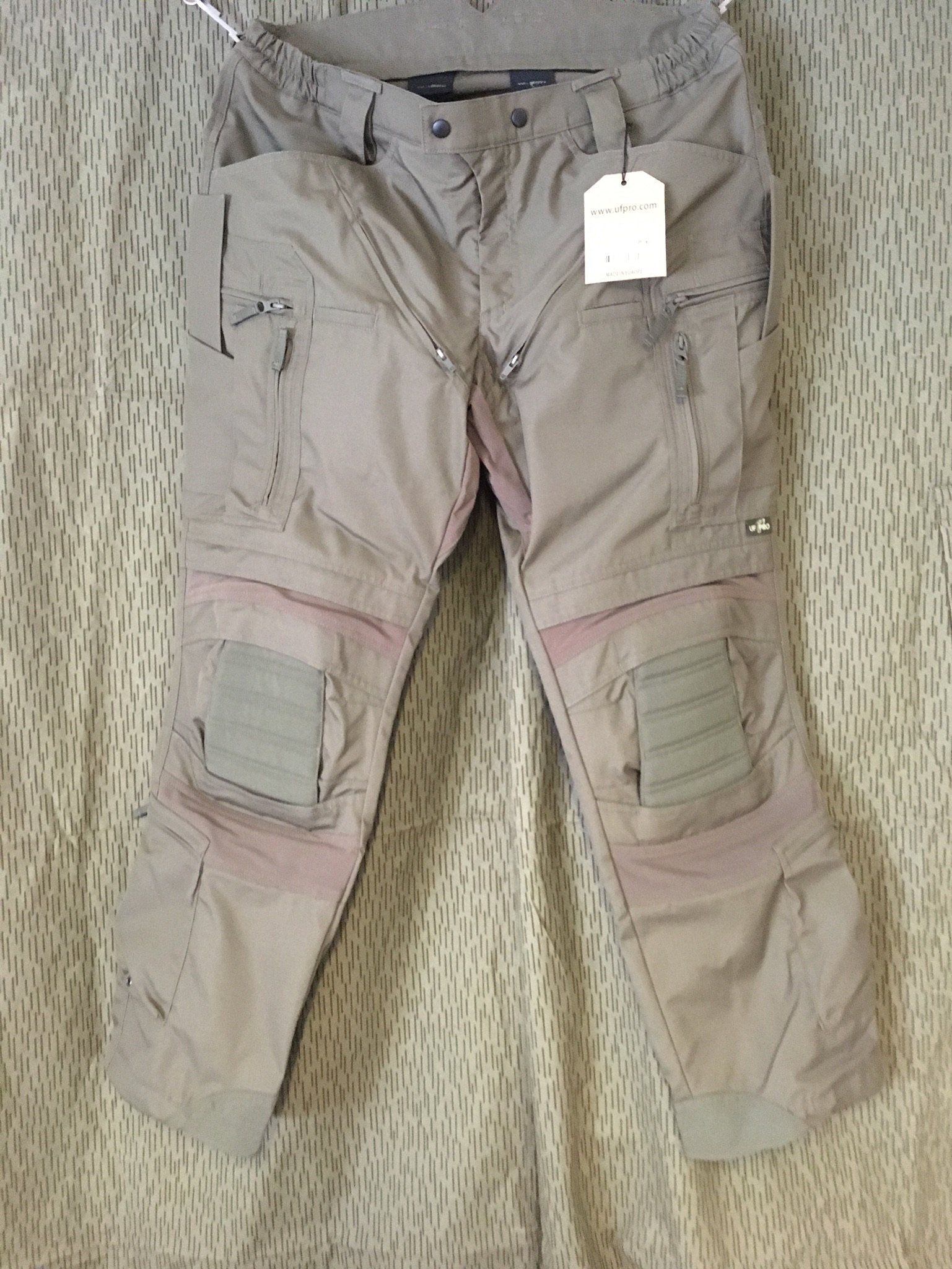 Hellbraune Cargohosen mit Knieflicken hängen vor einem Hintergrund mit Camouflage-Muster.