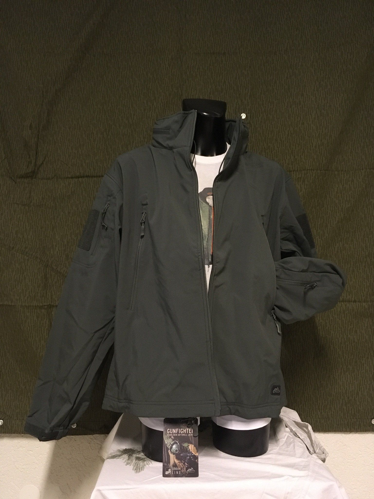 Dunkelgrüne Jacke an einer Schaufensterpuppe, mit Kapuze und Vordertasche. Vor einem Tarnhintergrund.