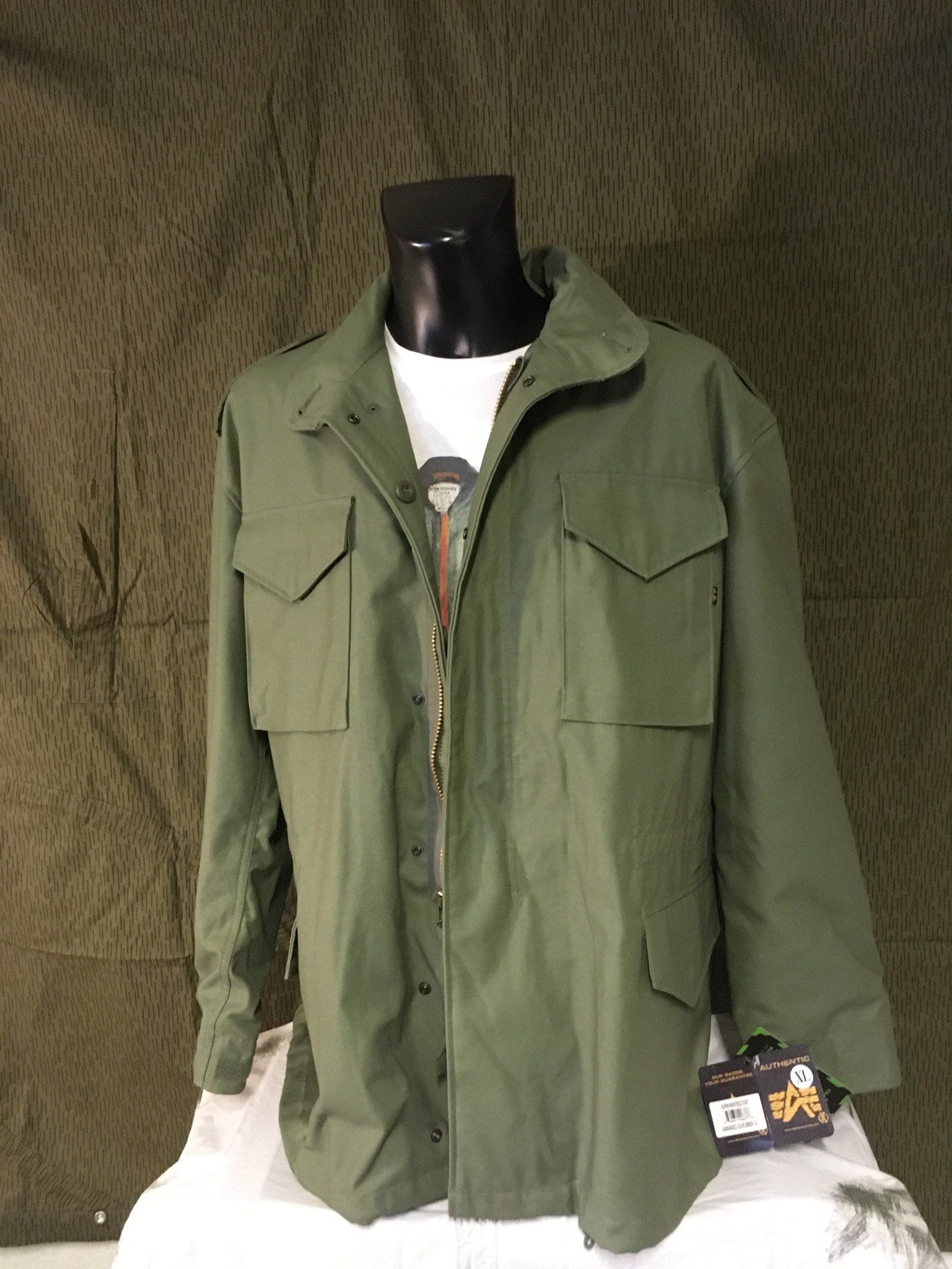 Grüne Jacke im Militärstil an einer schwarzen Schaufensterpuppe, über einem weißen T-Shirt, vor einem Tarnhintergrund.