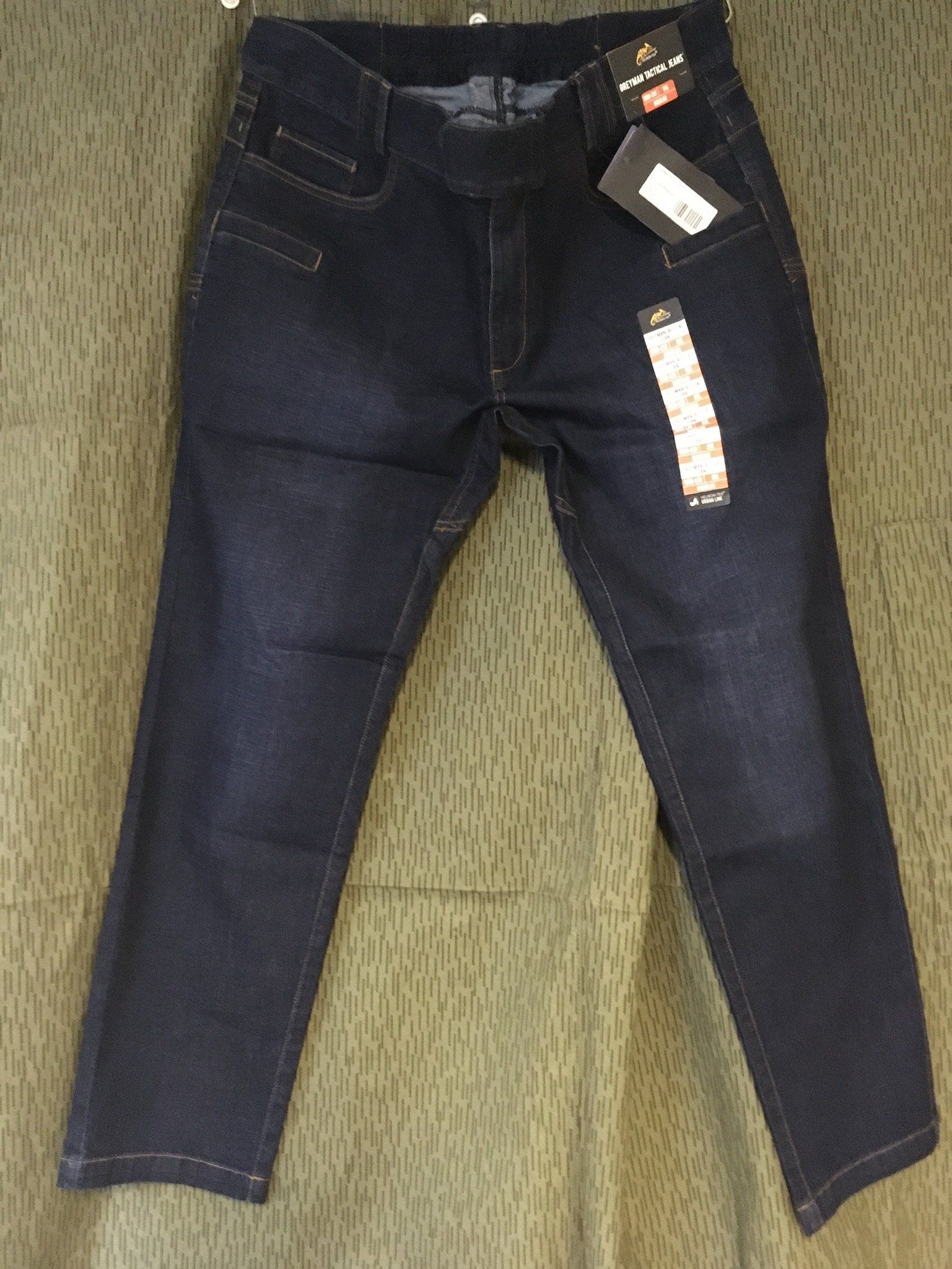 Dunkelblaue Jeans mit Etikett, die vor einem gemusterten Hintergrund hängt.