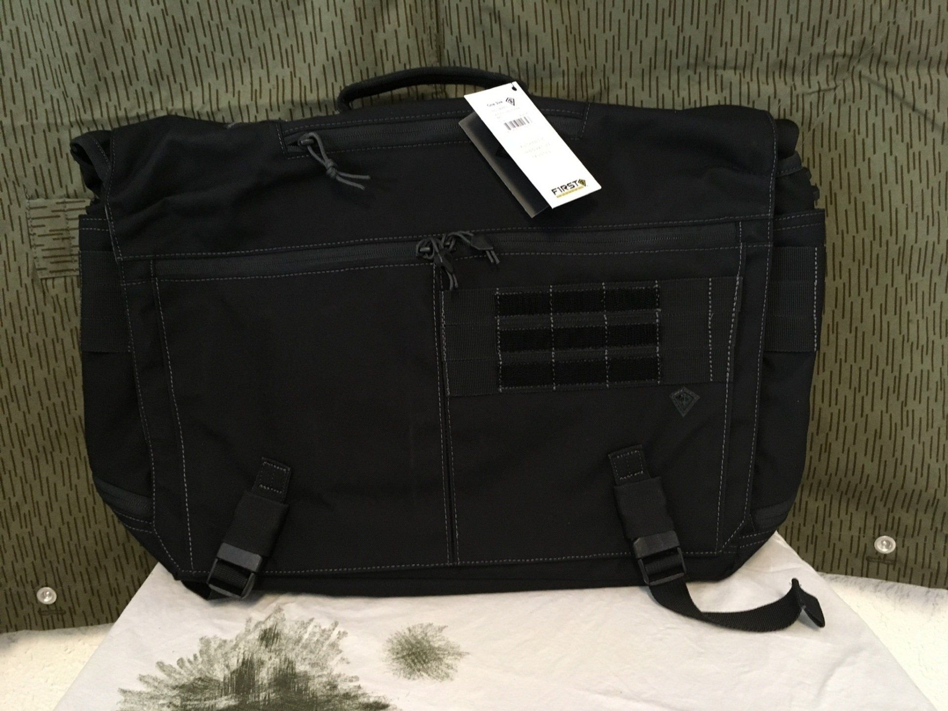 Schwarze Umhängetasche mit einem Etikett auf einer weißen Oberfläche vor einem Tarnhintergrund.