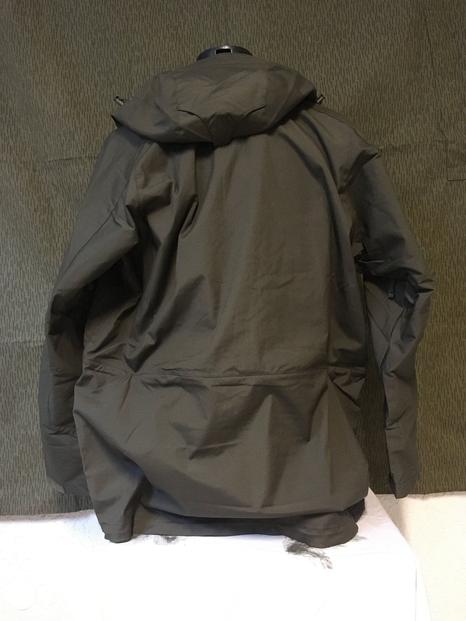 Rückansicht einer dunkelgrünen Kapuzenjacke, die an einer strukturierten Wand hängt.