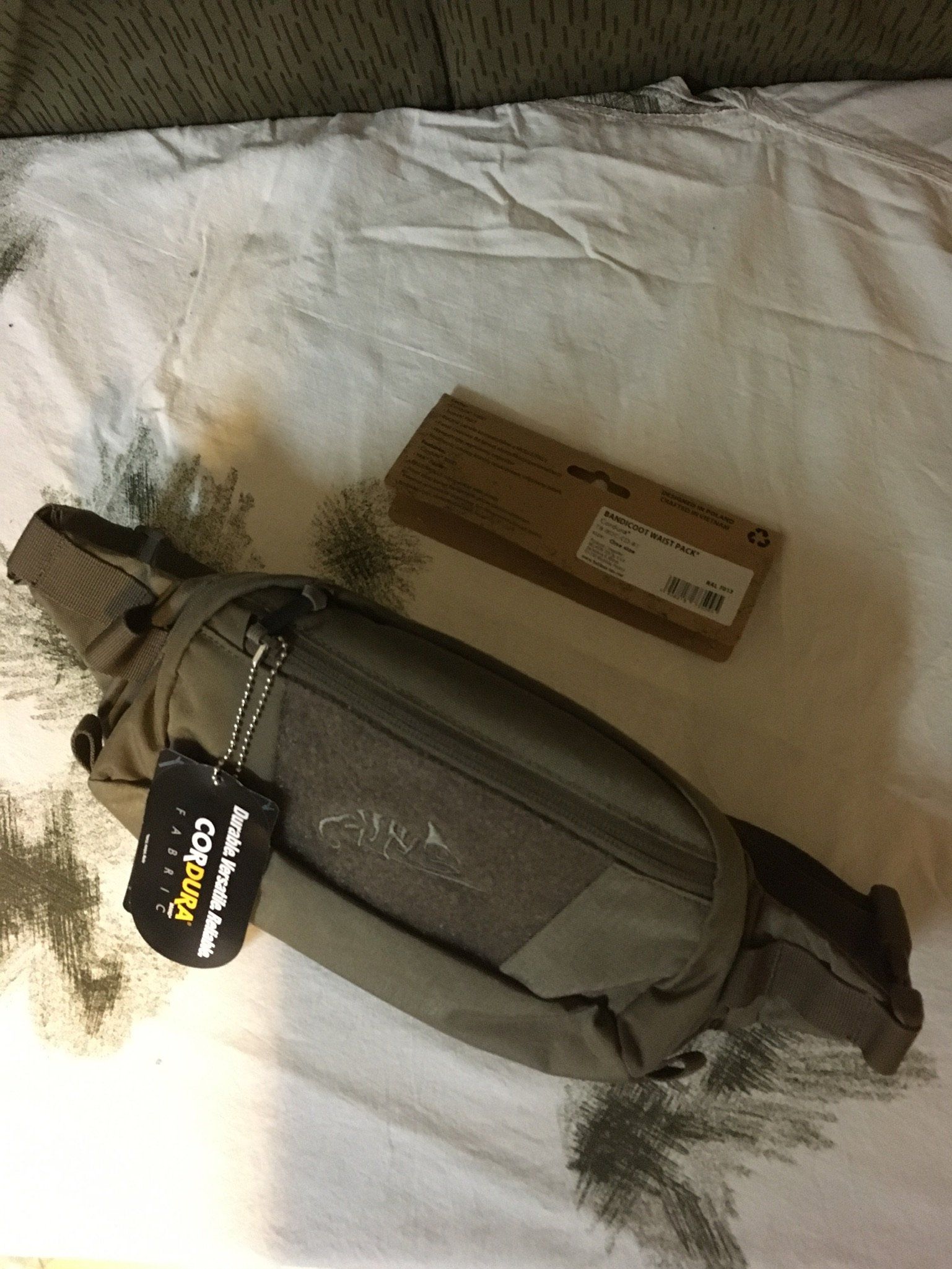 Gürteltasche und Karton auf einer zerknitterten weißen Oberfläche. Die Gürteltasche ist olivgrün und mit einem Etikett versehen.