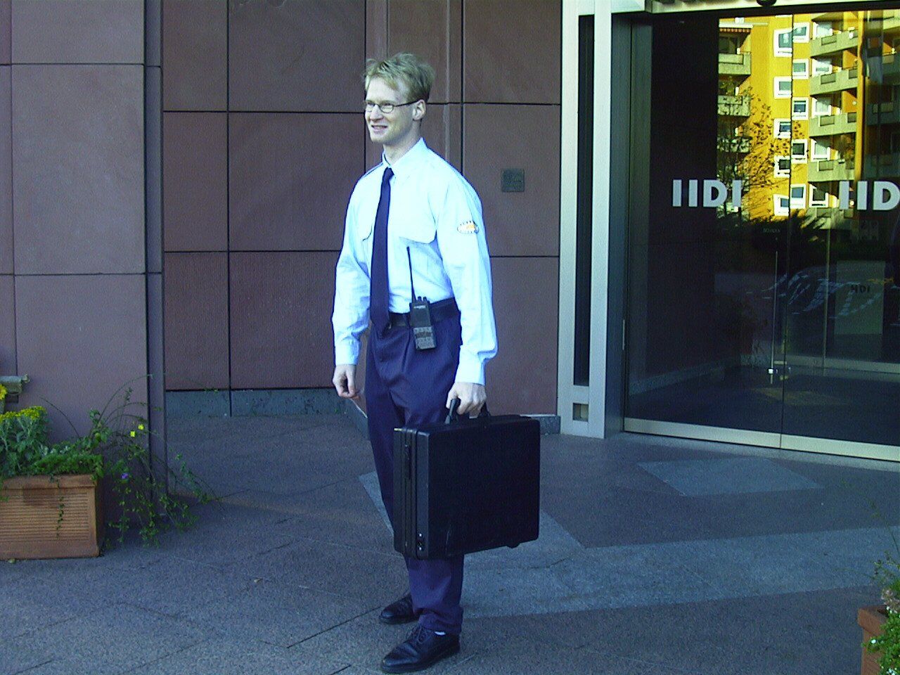 Mann in Geschäftskleidung vor einem Gebäude, hält Aktentasche in der Hand und lächelt.