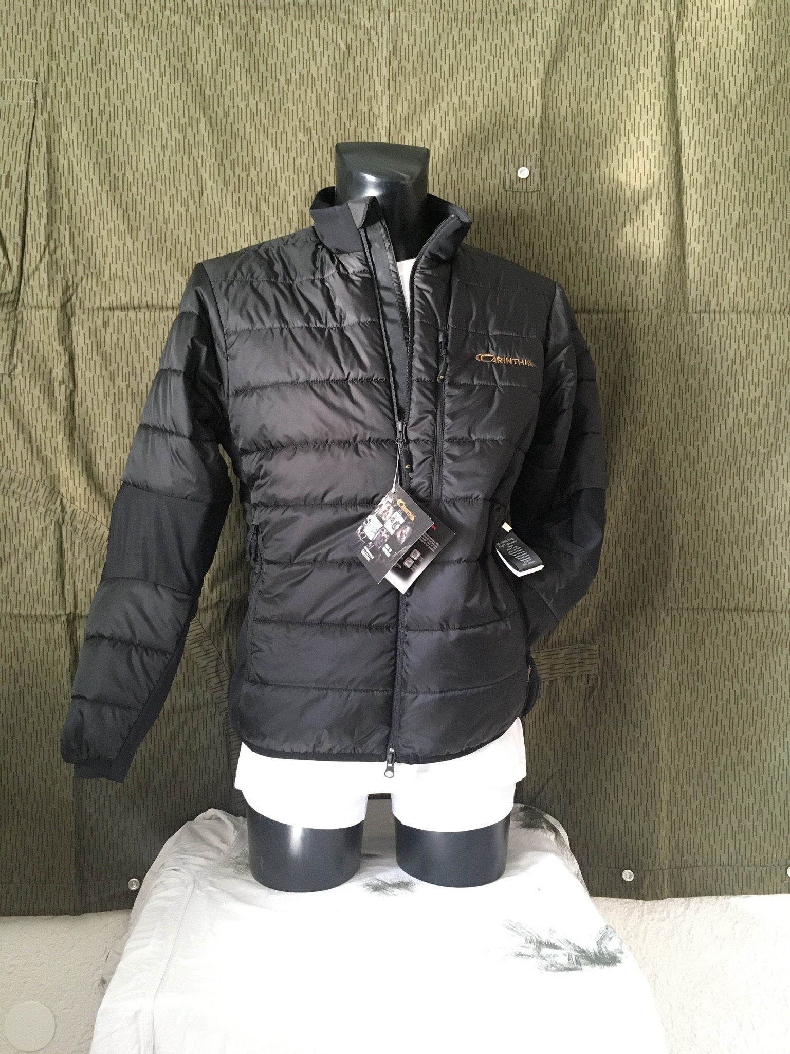 Schwarze Pufferjacke an einer Schaufensterpuppe, mit angebrachten Etiketten, vor einem Tarnhintergrund.