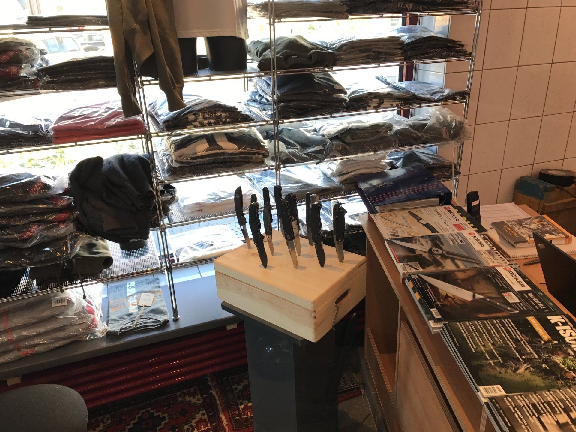 Messer in einem Holzblock auf einem Ständer, neben einem Zeitschriftenständer und Regalen mit gefalteter Kleidung.