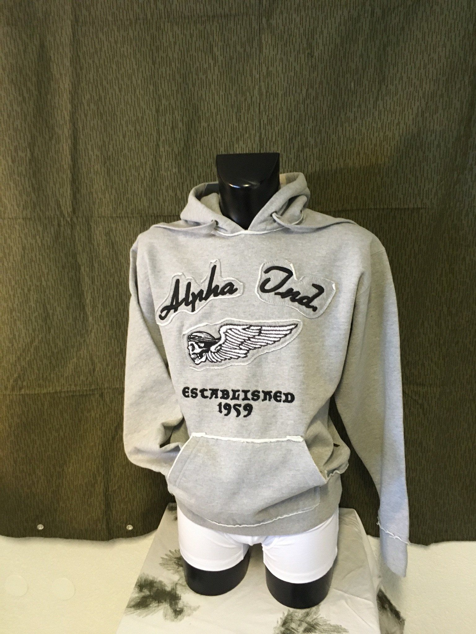 Grauer Kapuzenpullover an einer Schaufensterpuppe mit „Alpha One“-Logo und Tasche. Gegründet 1969. Vor einem melierten grünen Hintergrund.