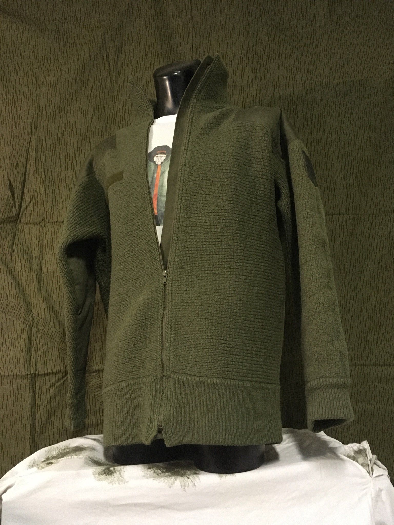 Olivgrüne Jacke auf einer Schaufensterpuppe, Vorderansicht. Mit Reißverschluss und hohem Kragen. Hintergrund: gemusterter grüner Stoff.