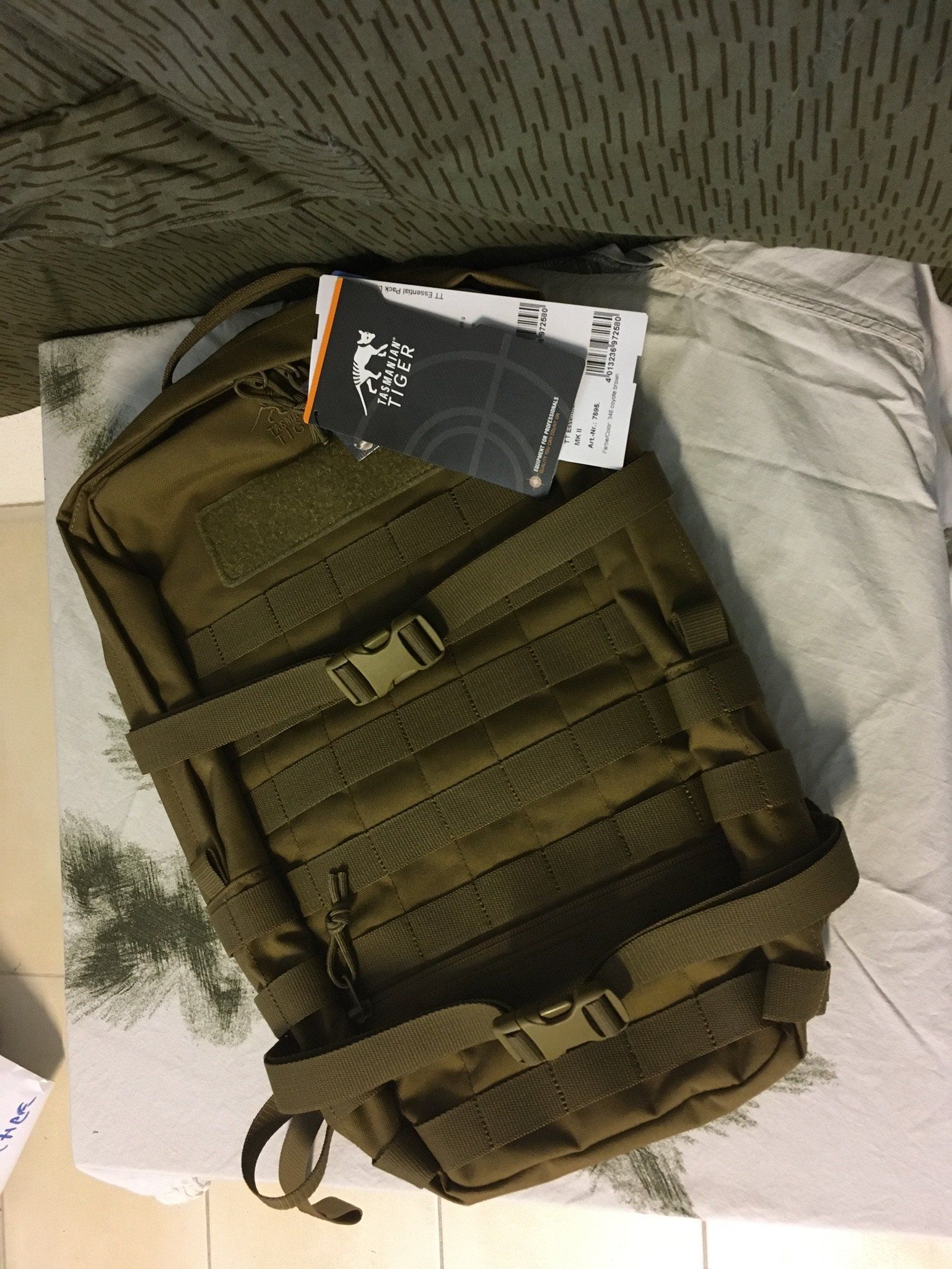 Hellbrauner Rucksack mit mehreren Riemen und Schnallen auf einer weiß-grünen Oberfläche.