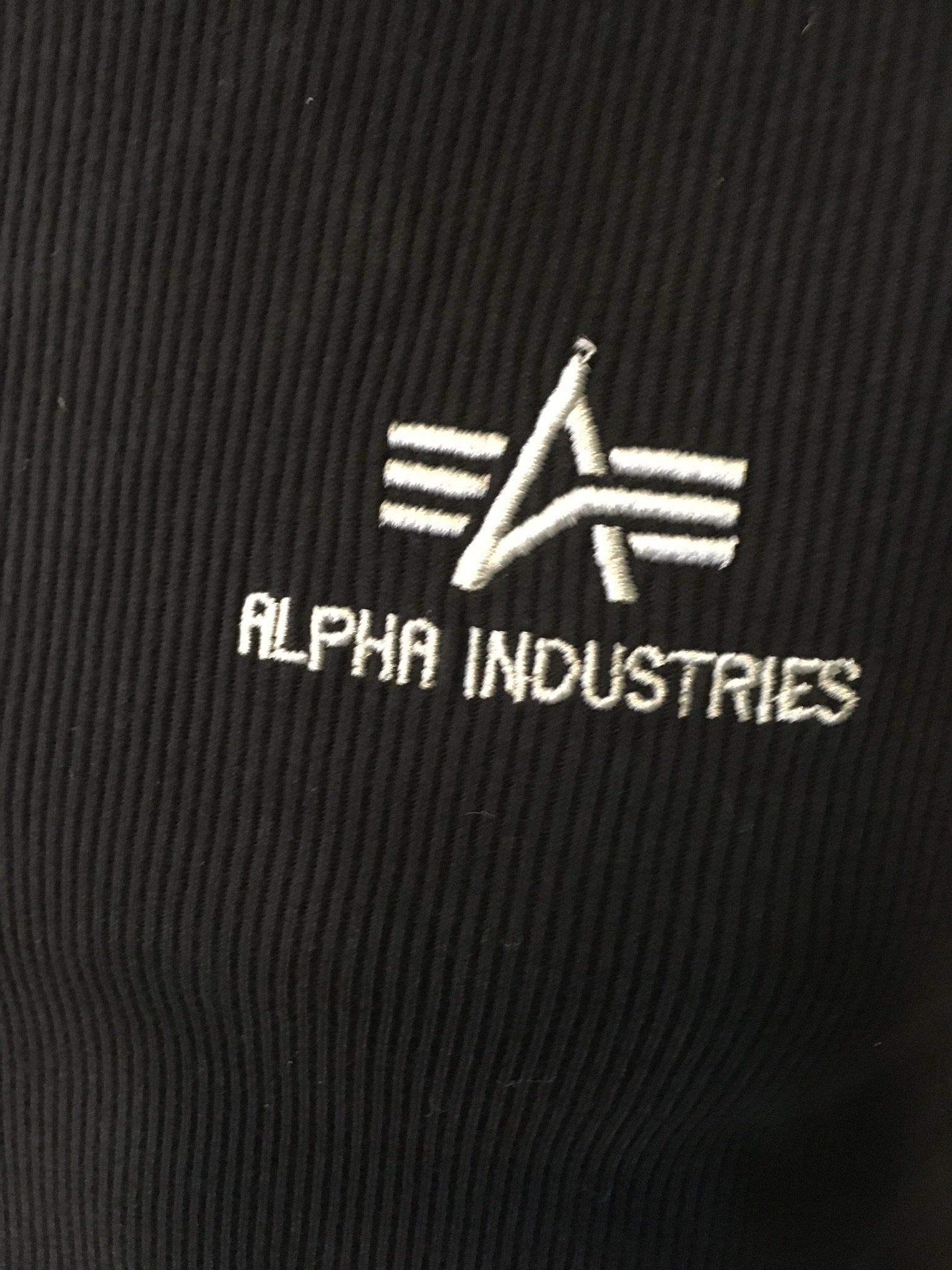 Gesticktes Alpha Industries-Logo in Weiß auf schwarzem Stoff.