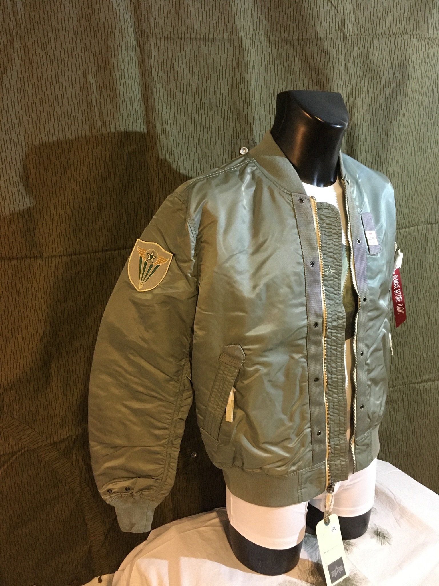 Olivgrüne Bomberjacke an einer Schaufensterpuppe, mit Flicken und einem sichtbaren weißen Hemd.