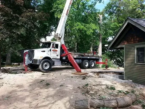 Crane truck removing tree near a house; beige, white, green; outdoor setting.