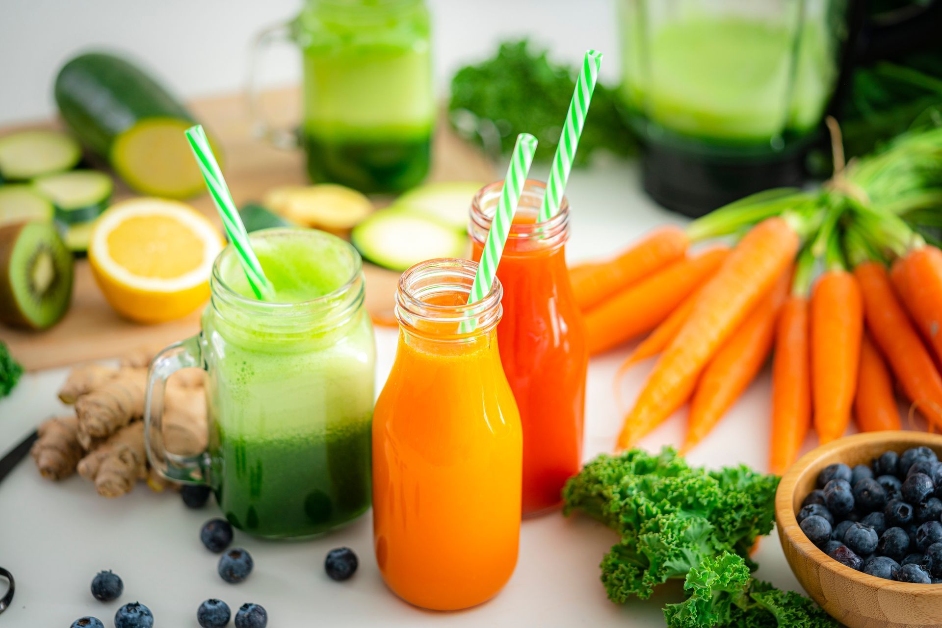 Three smoothies in mason jars with straws on a table surrounded by fruits and vegetables.