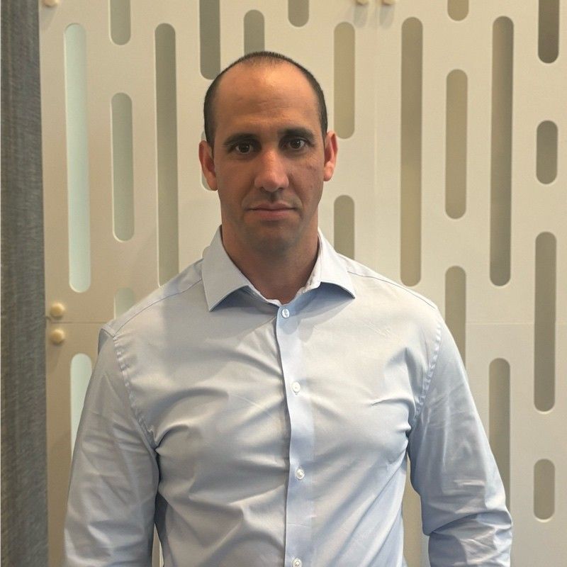 Man in a light blue button-down shirt stands in front of a white patterned wall.