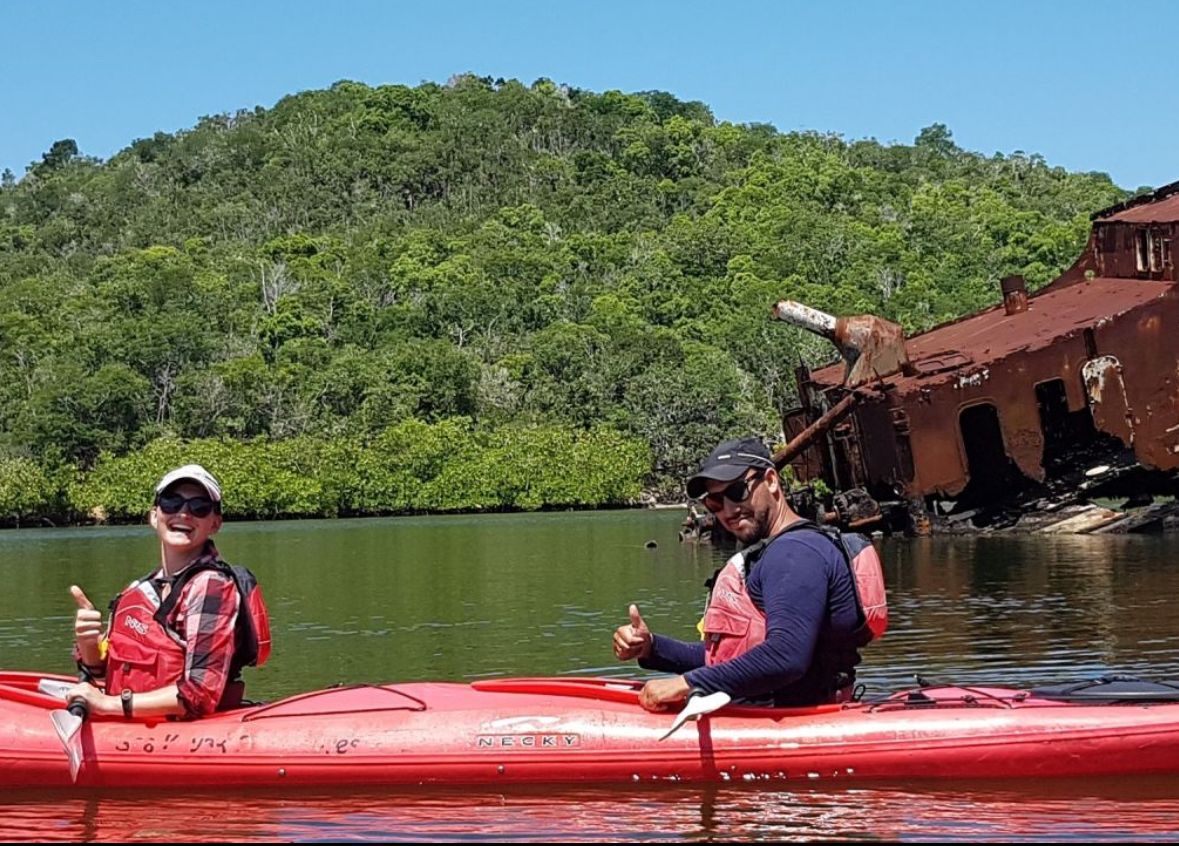 Two people kayaking, thumbs up, near a rusty shipwreck and green landscape.