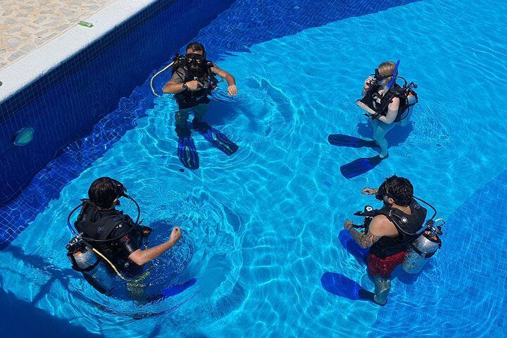 Four people in scuba gear in a bright blue pool, practicing.