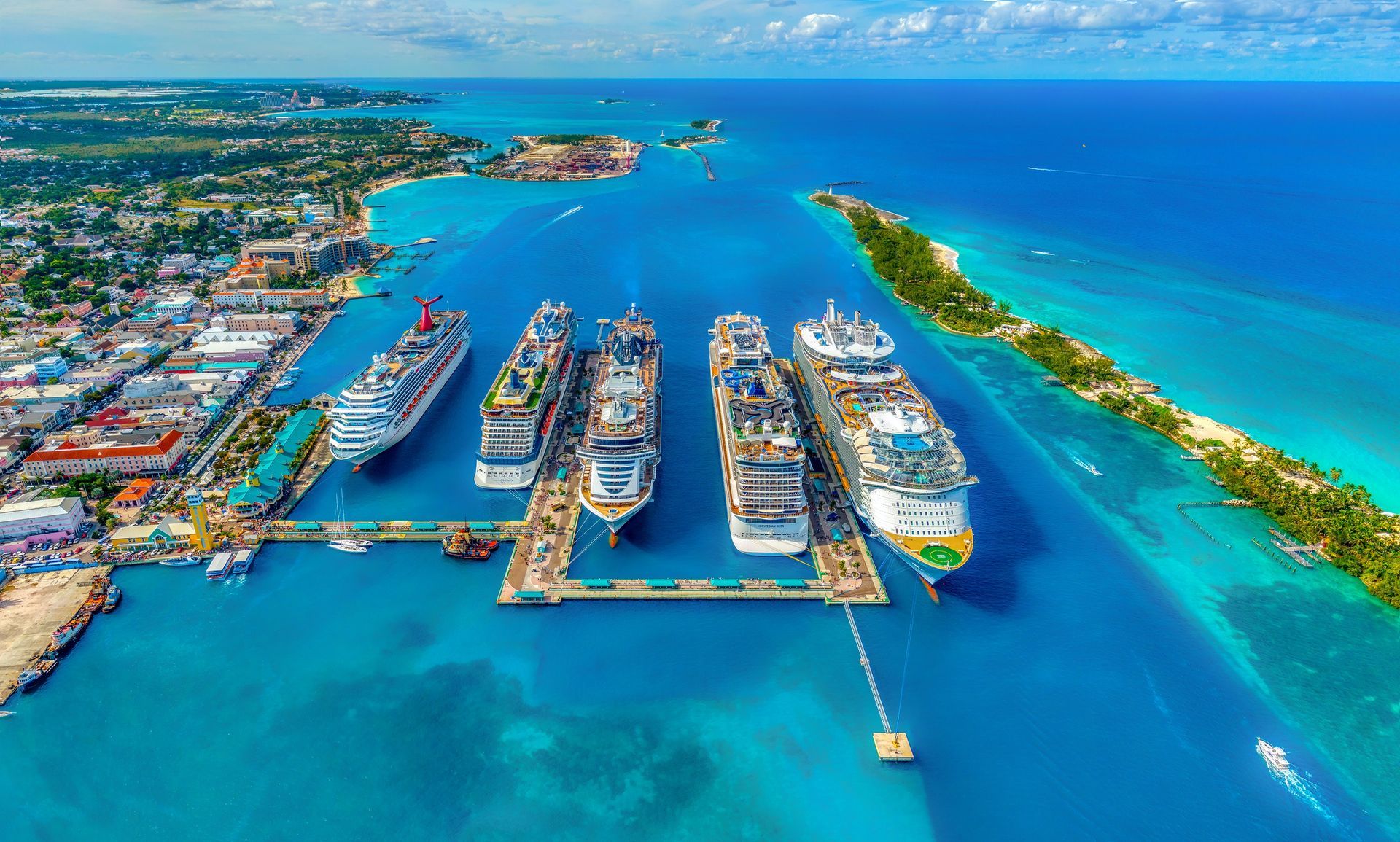 Cruise ships docked in a turquoise harbor, lined up along a pier, with buildings and a small island visible.