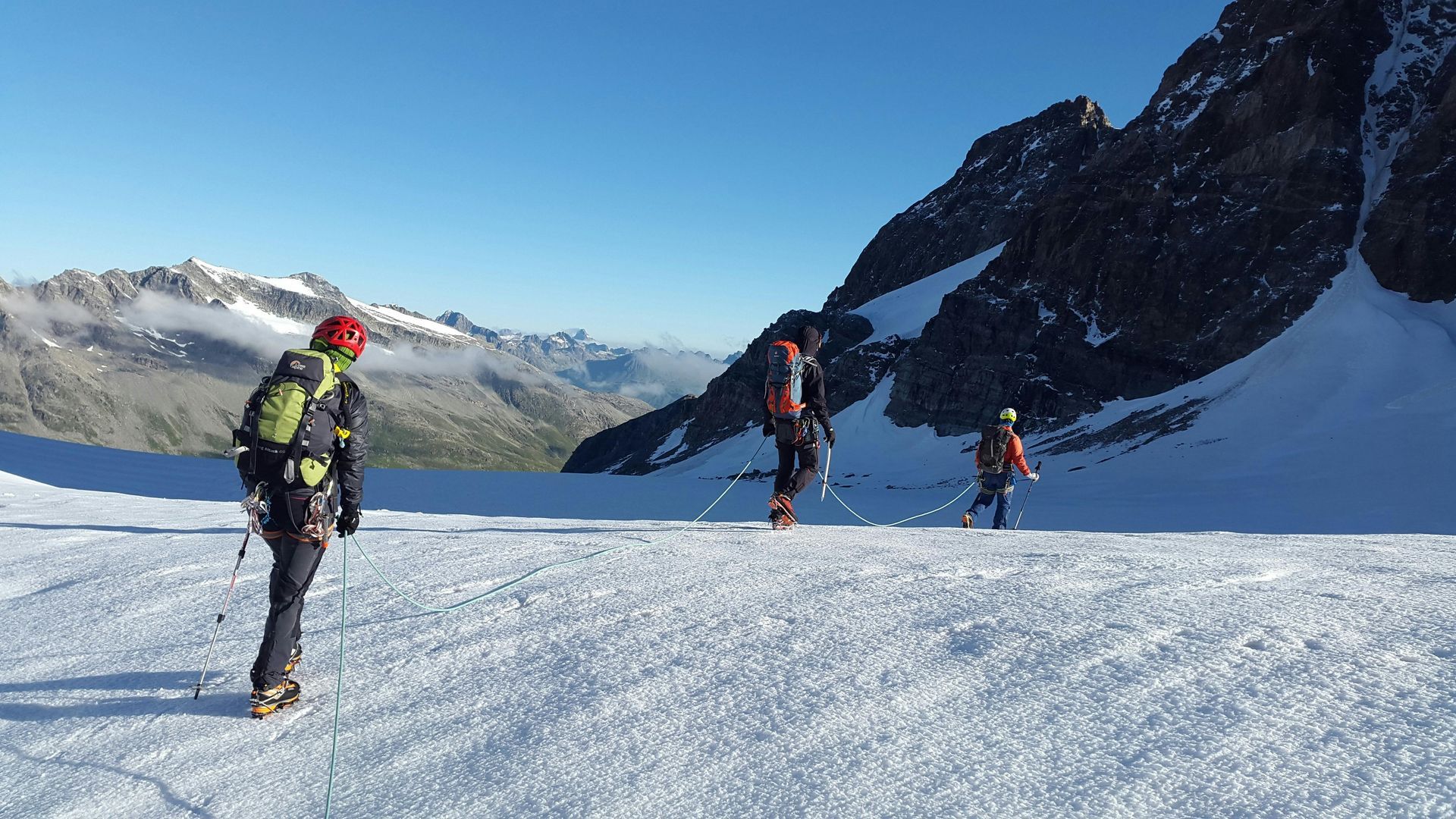 Three people on skis and with backpacks traversing a snow-covered mountain slope, with peaks in the background.