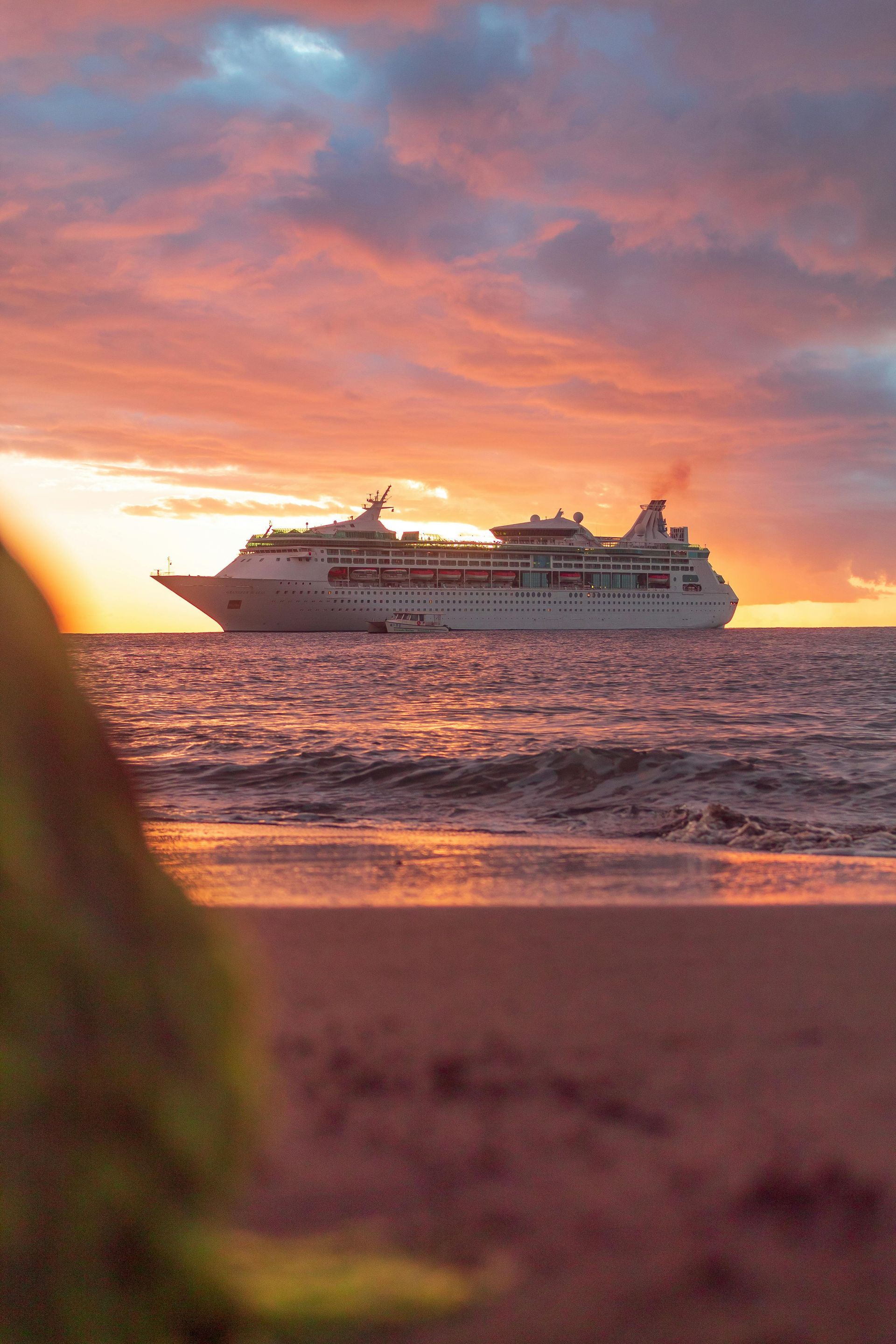 Cruise ship sailing on ocean at sunset, orange and pink sky above the water, beach in foreground.