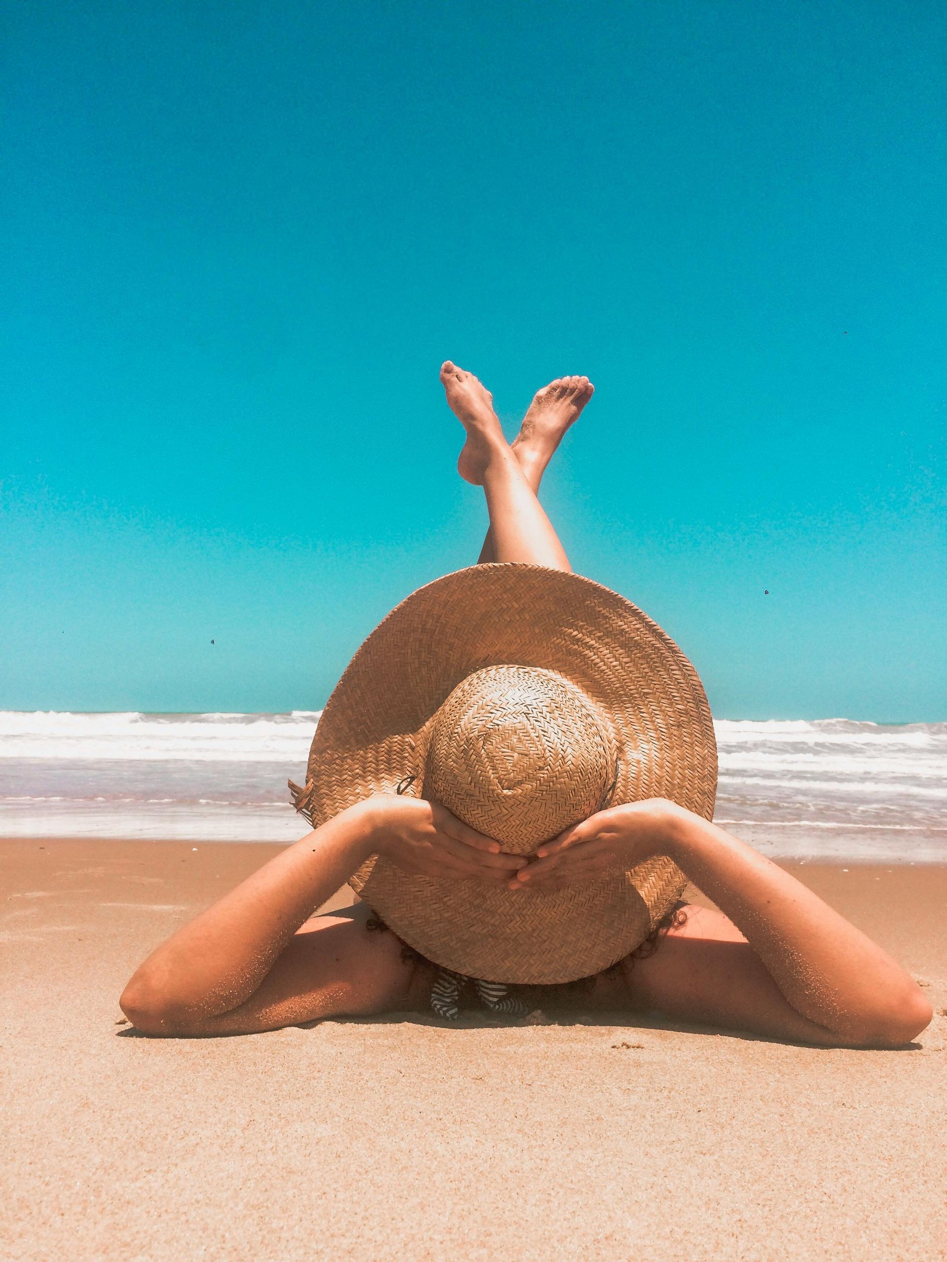 Person lying on beach, wearing large straw hat, arms near face, legs crossed, blue sky, ocean in background.