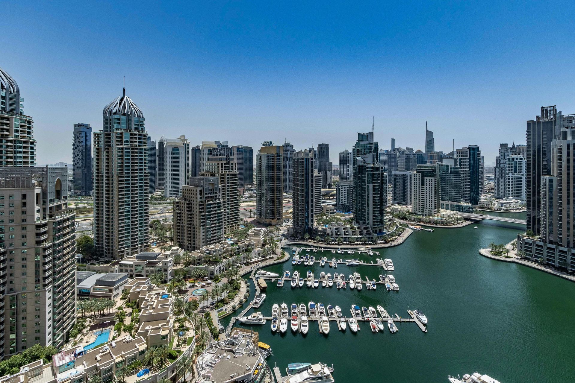 Dubai Marina cityscape, many tall modern buildings around a marina filled with docked boats, blue sky.