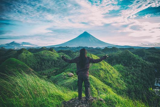 Person with arms outstretched, overlooking green hills and a mountain under a blue sky.