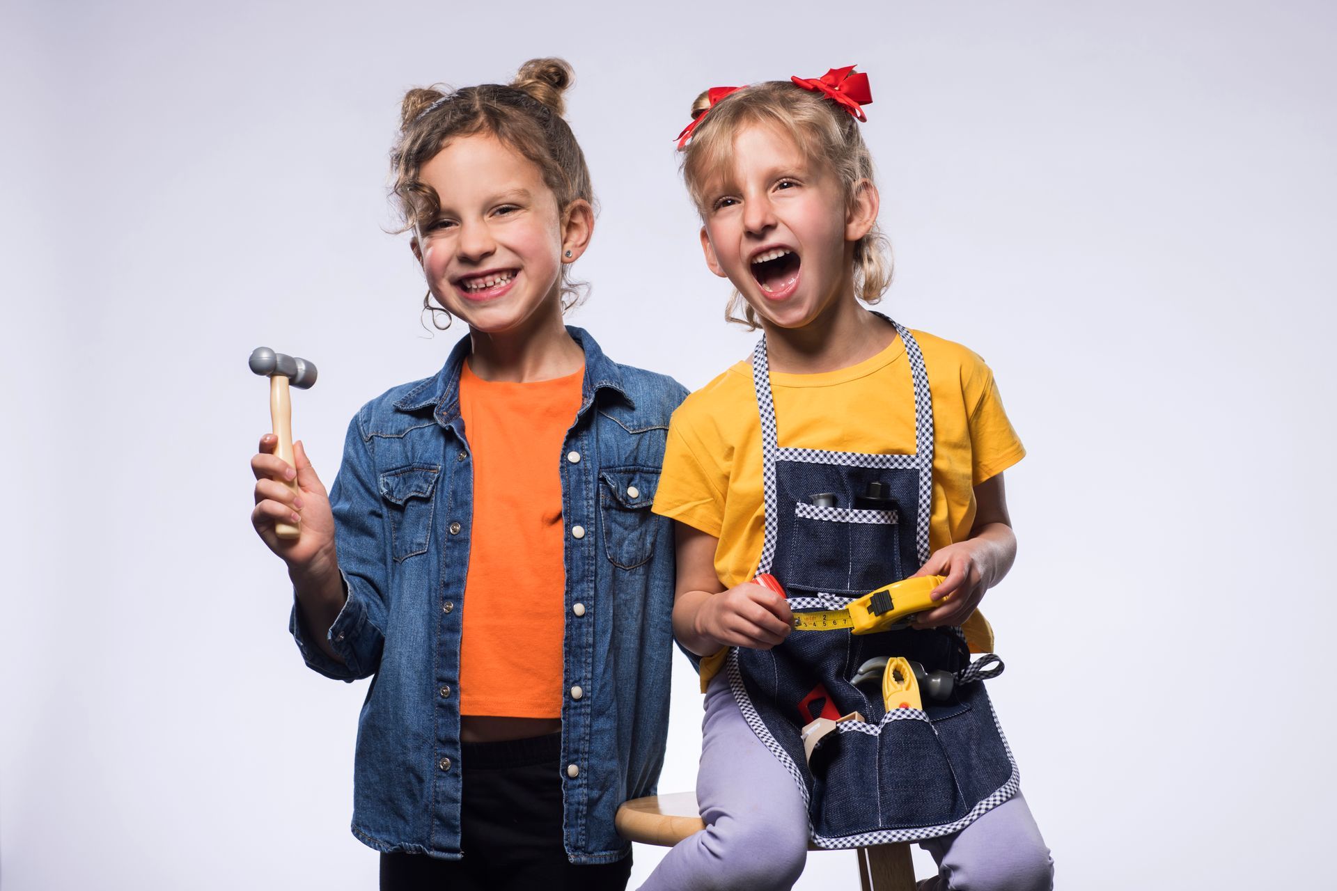 Two girls with tools, one holding a hammer, smiling. The other, wearing a tool belt, is laughing.