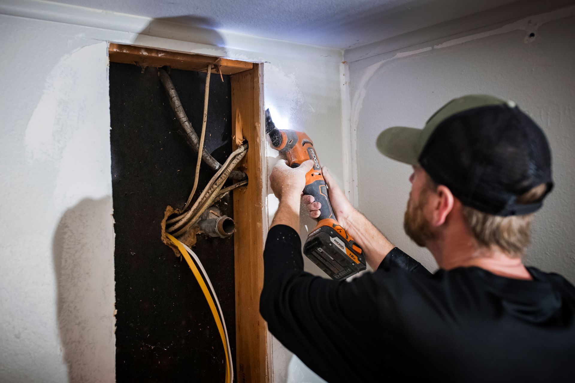 Man using a reciprocating saw to cut into a wall, revealing wires and plumbing. Indoors, bright light.