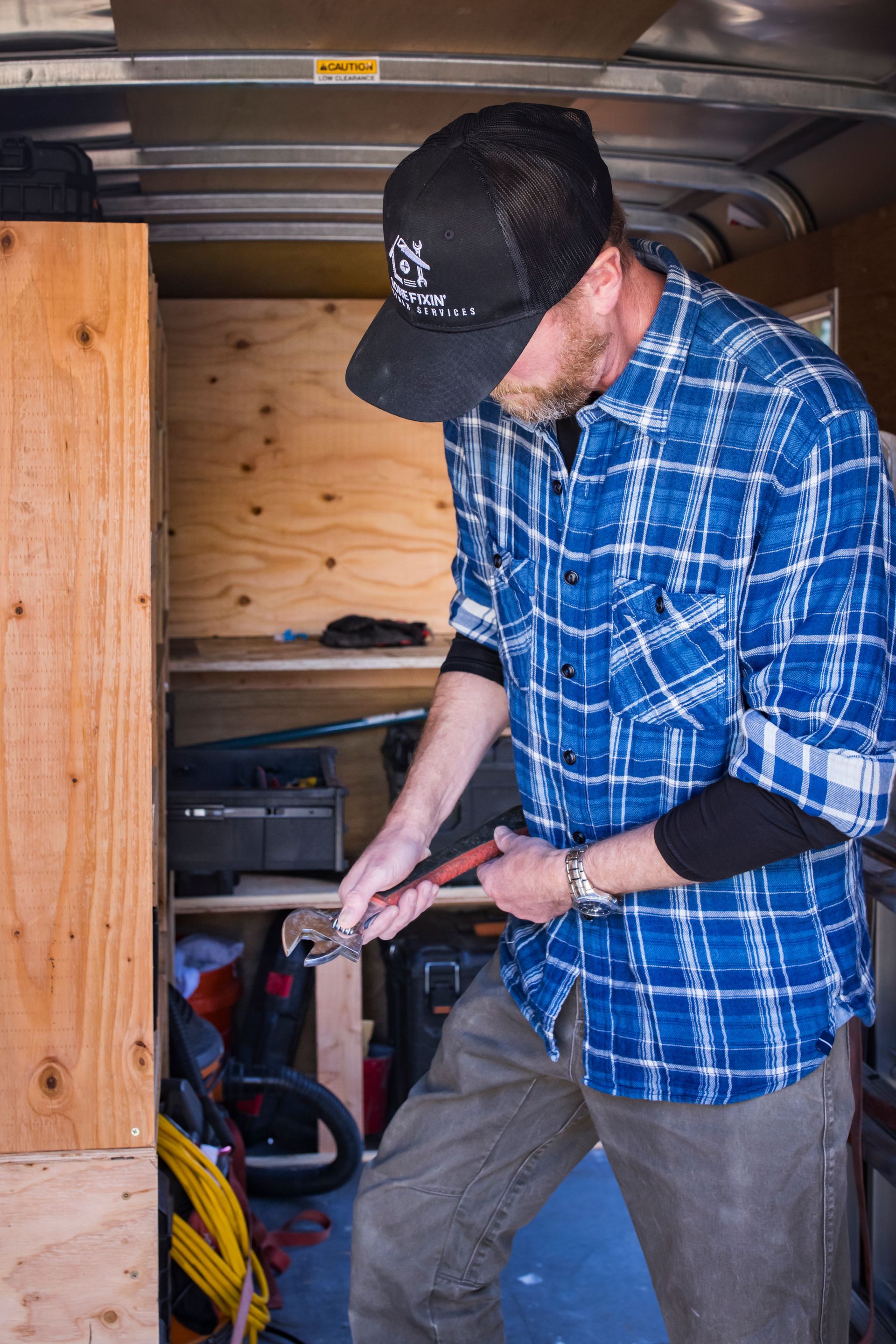 Man in blue plaid shirt and cap using a tool inside a wooden storage area.