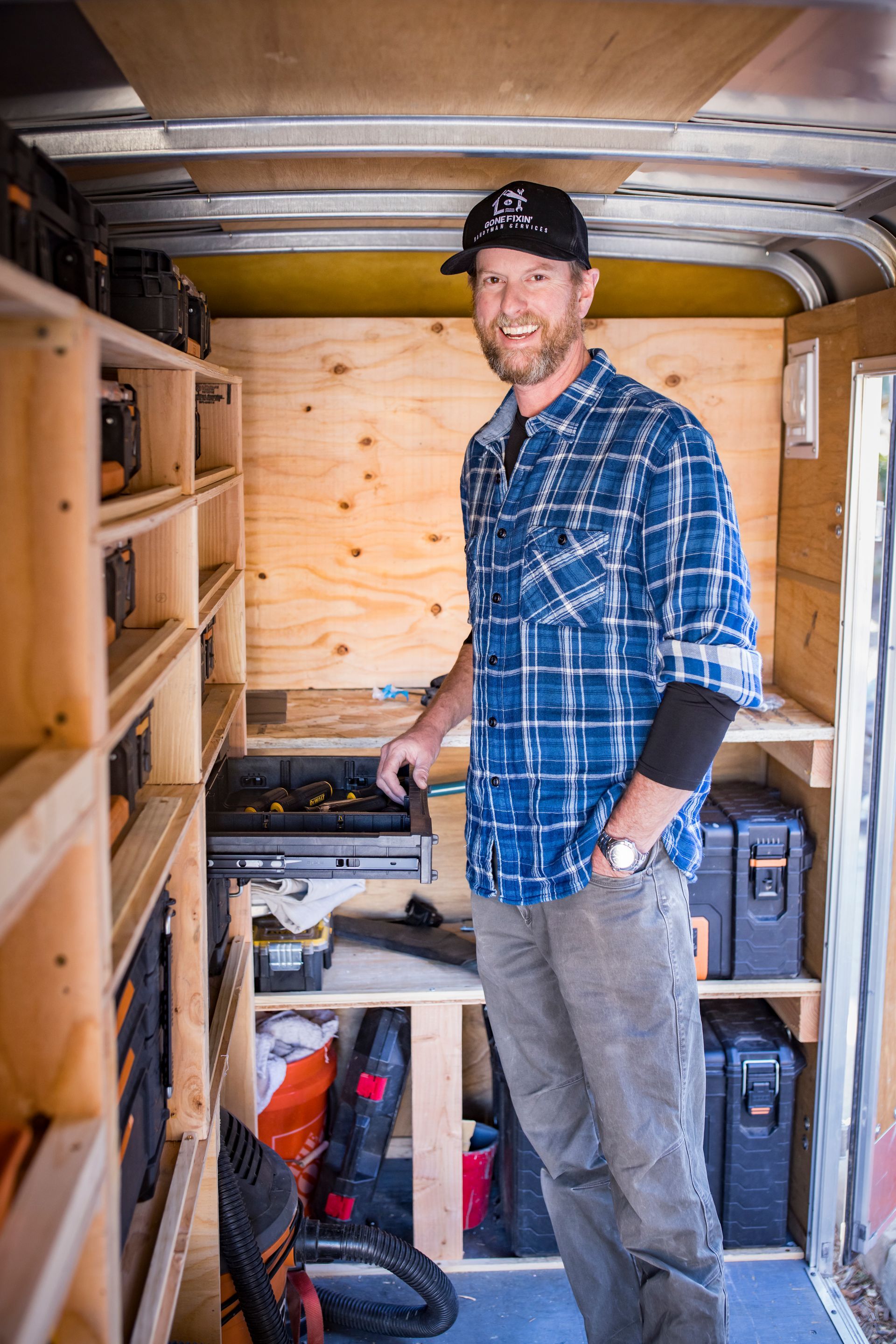 Man in plaid shirt standing in a trailer with wooden shelving, smiling and holding a tool.