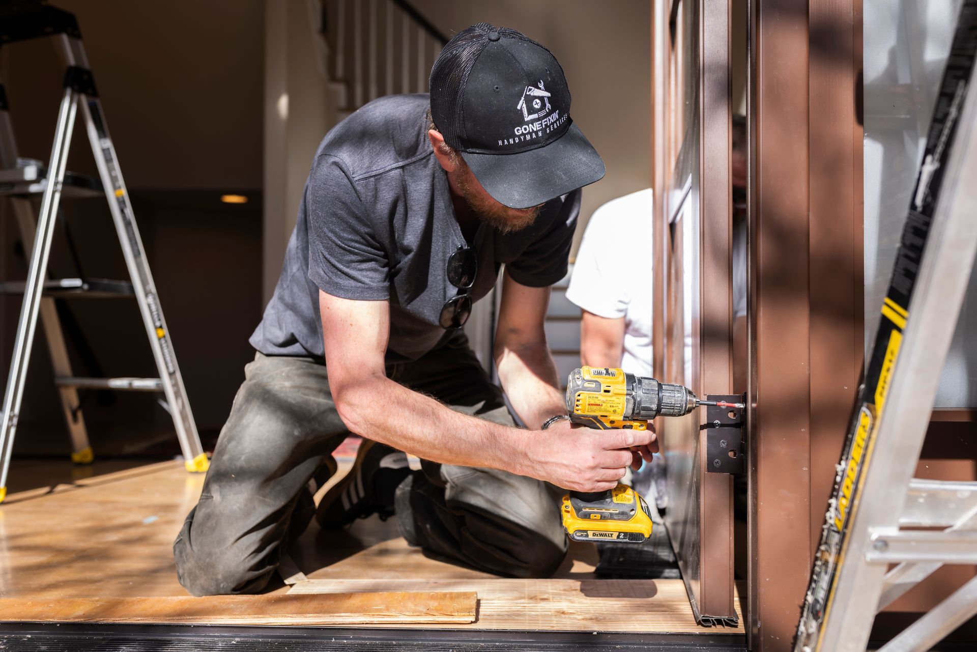 Man using a power drill on a door hinge, another person assisting. Wooden door frame, interior setting.