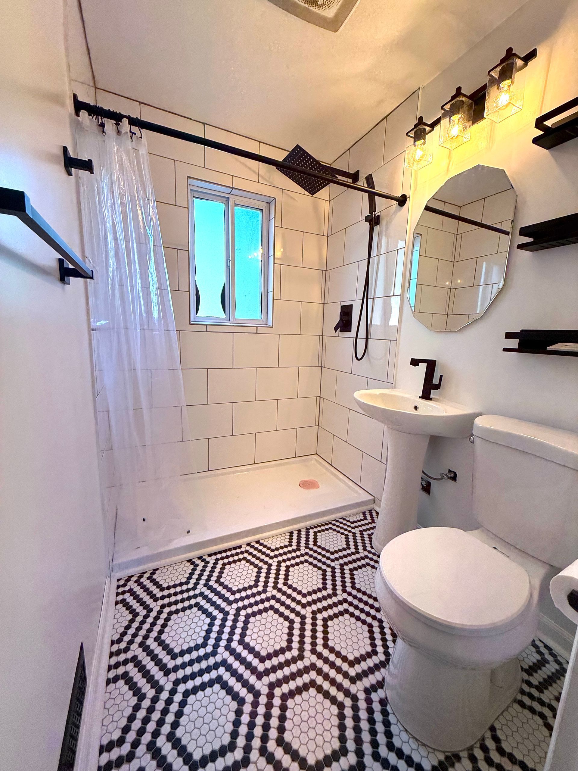 Small, modern bathroom with patterned floor, white tiles, shower, toilet, pedestal sink, and black fixtures.