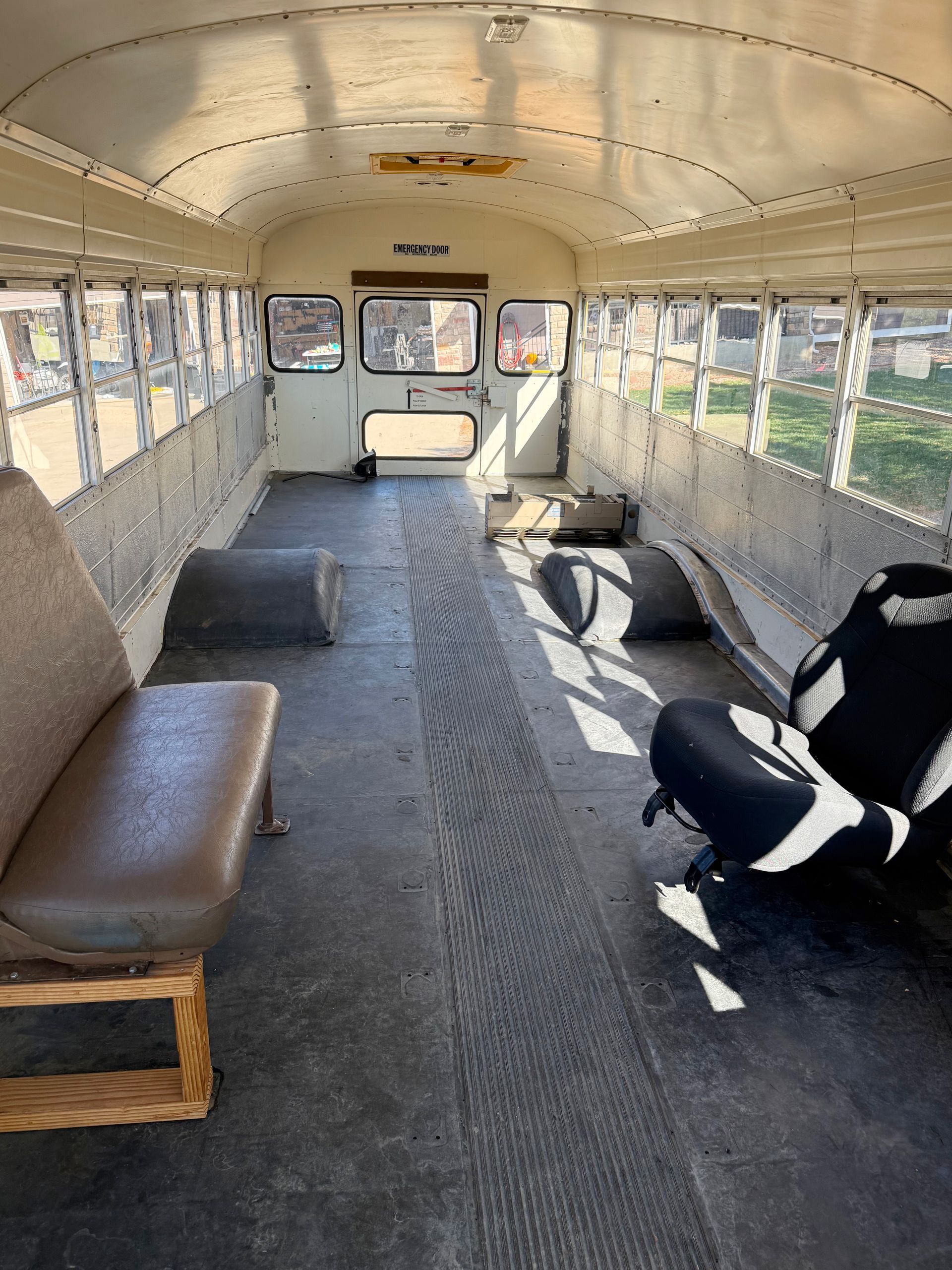 Interior of a school bus with seats removed. Beige walls, dark floor. Daylight streams through windows.