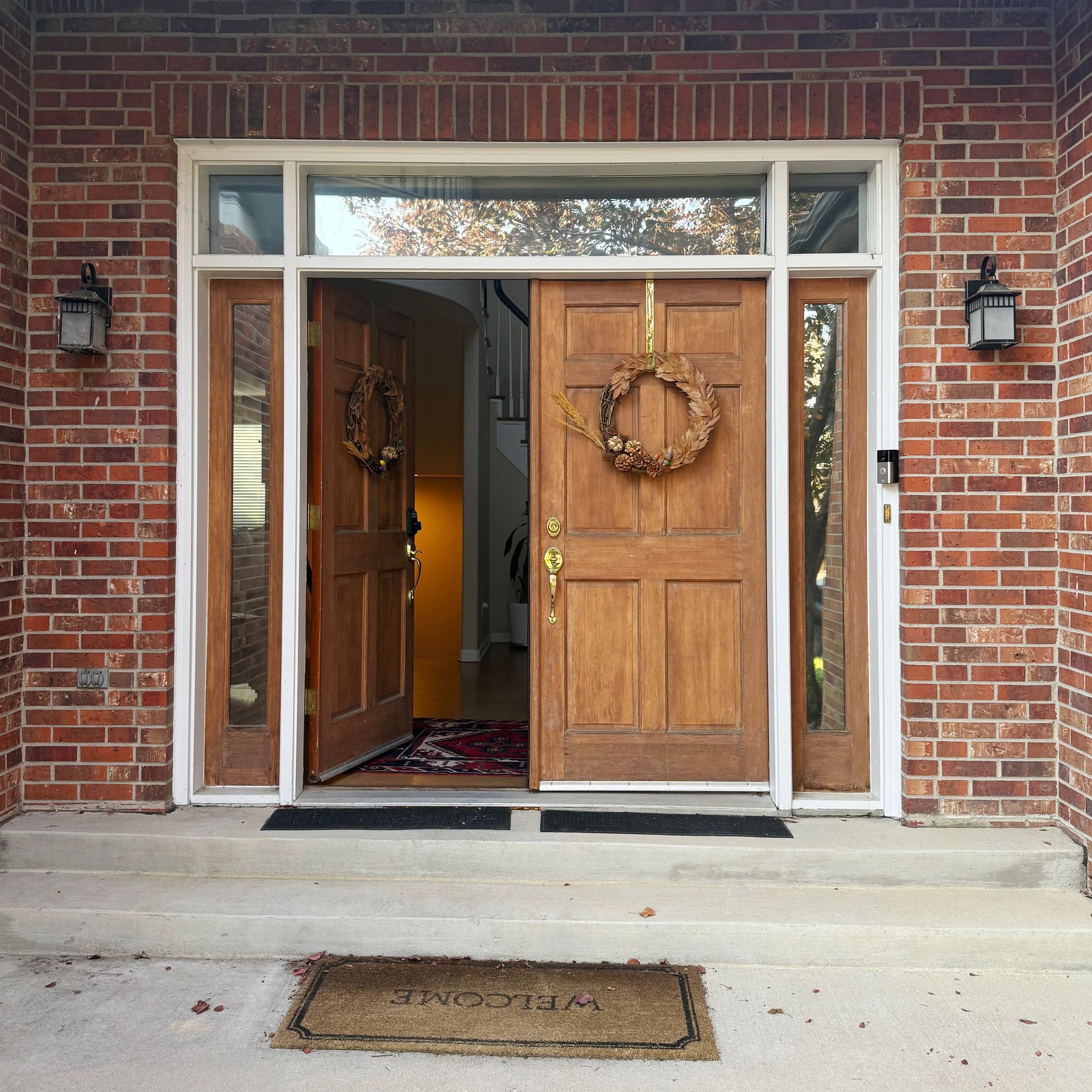 Brick building entrance with open wooden double doors, wreaths, sidelights, and overhead transom.