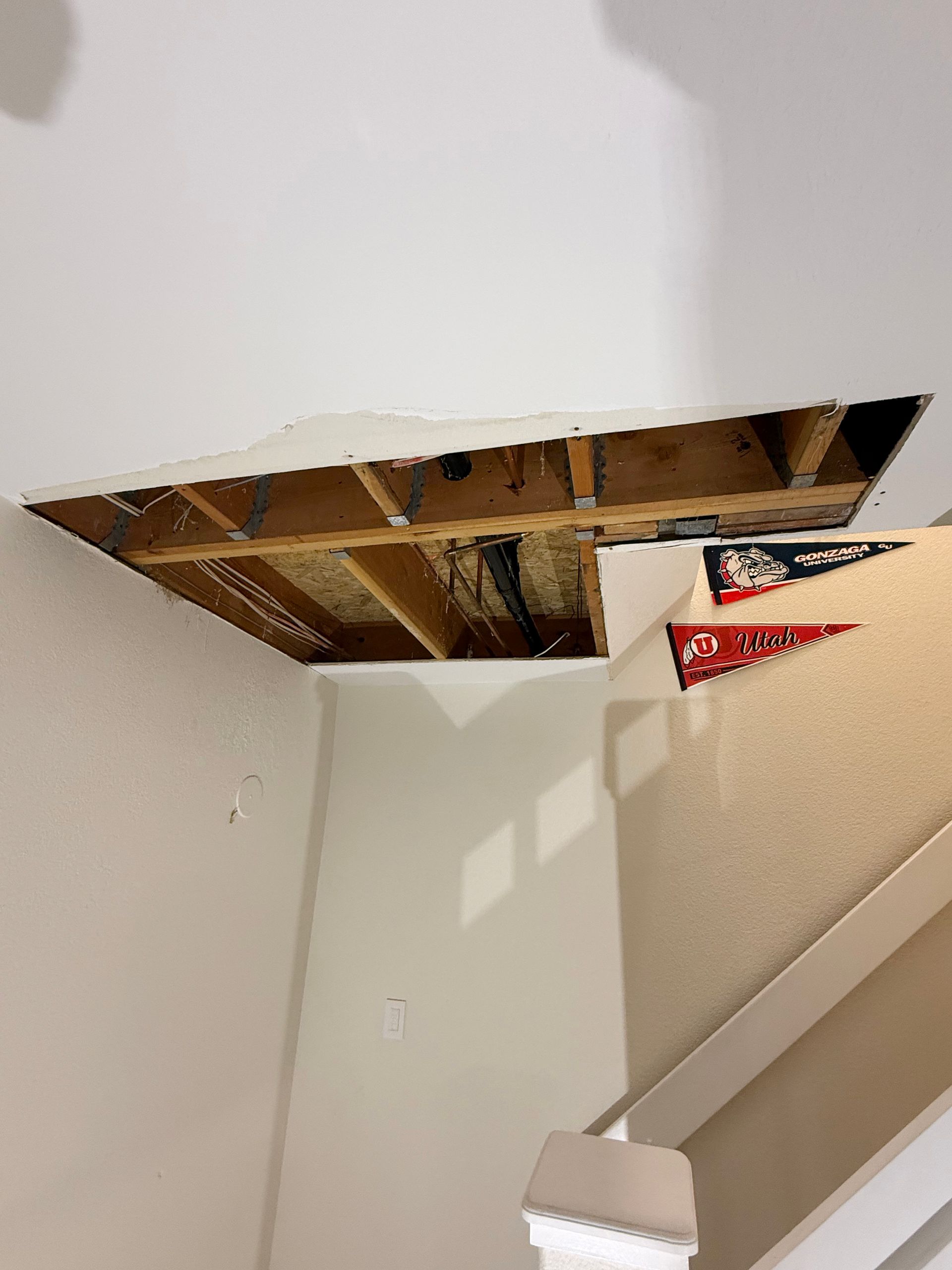 A rectangular hole in a white ceiling reveals exposed wooden joists, wiring, and plumbing pipes above a staircase.