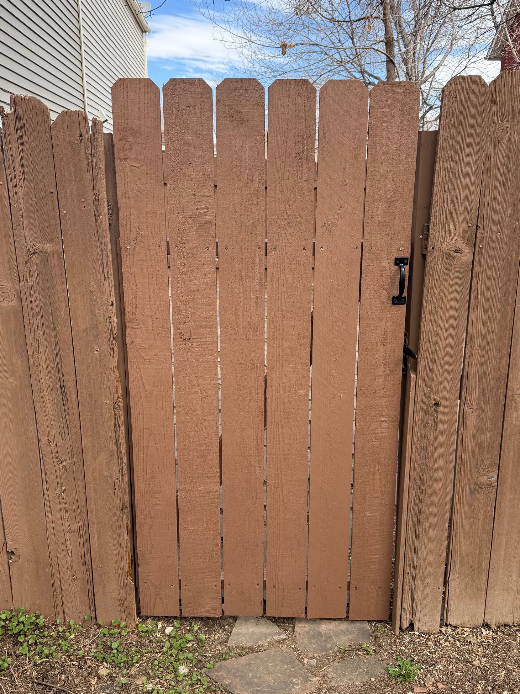 A brown wooden picket gate set within a matching fence, with a black metal handle on the right side.