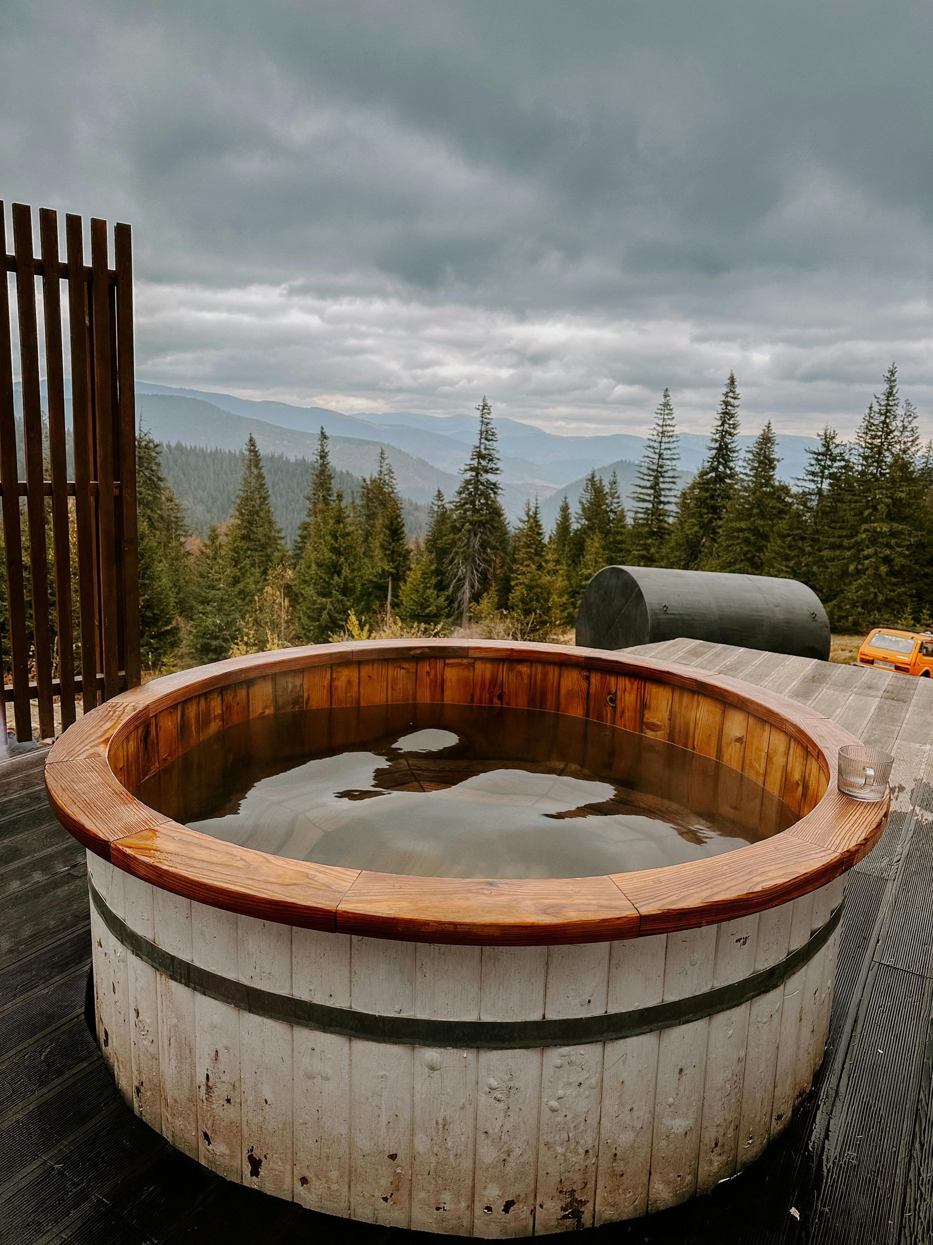 Wooden hot tub filled with water on a deck overlooking a mountain range under a cloudy sky.