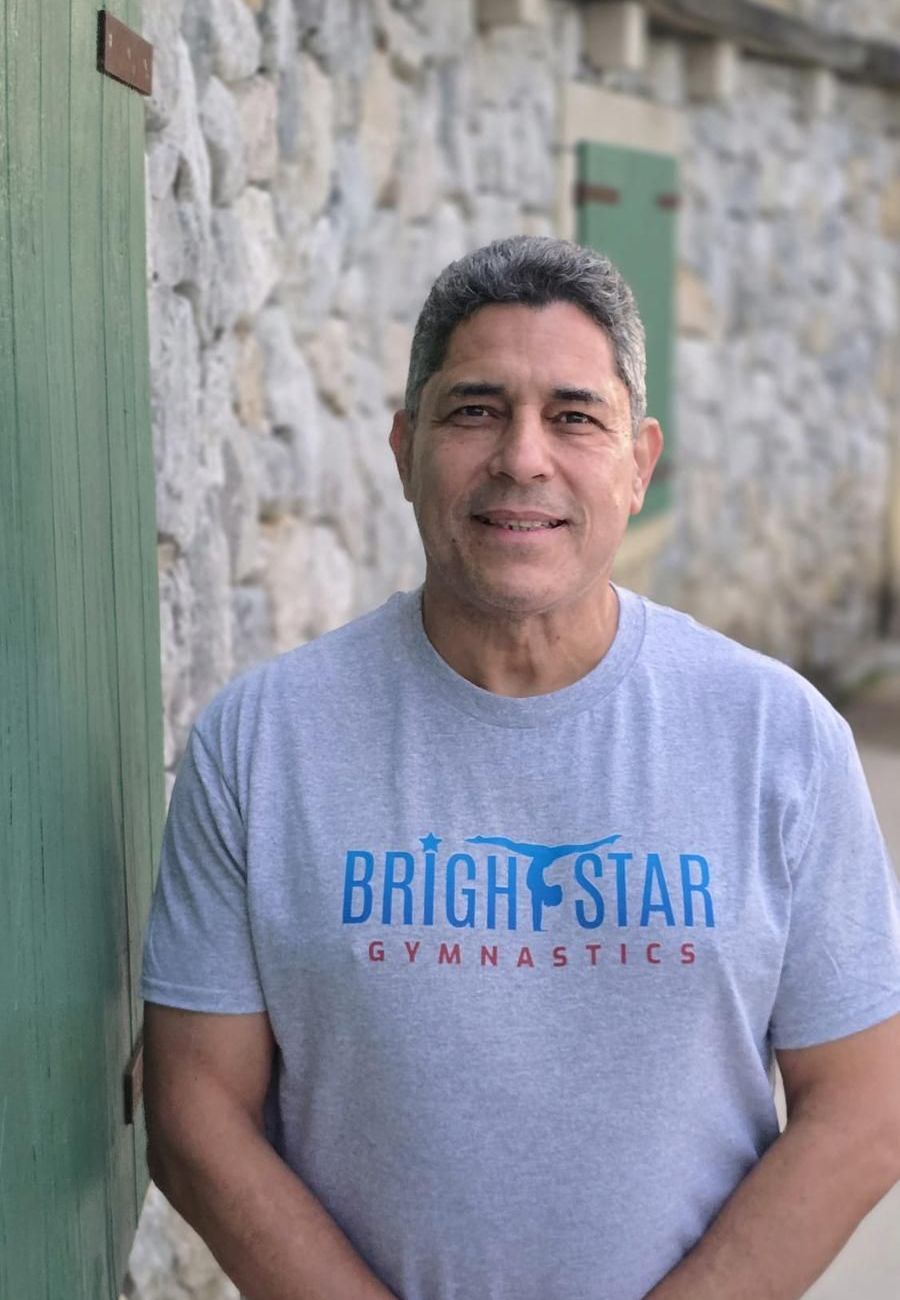 A man wearing a bright star gymnastics t-shirt is standing in front of a stone wall.