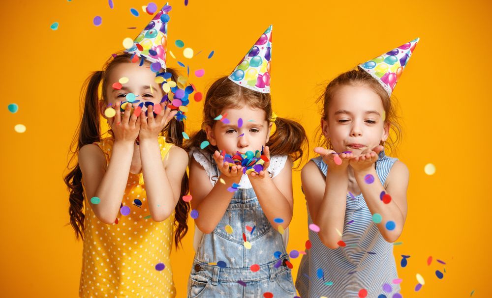 Three little girls wearing party hats are blowing confetti in the air.