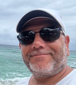 Un hombre con barba lleva gafas de sol y un sombrero en la playa.