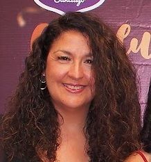 Una mujer con cabello largo y rizado sonríe frente a una pared violeta.