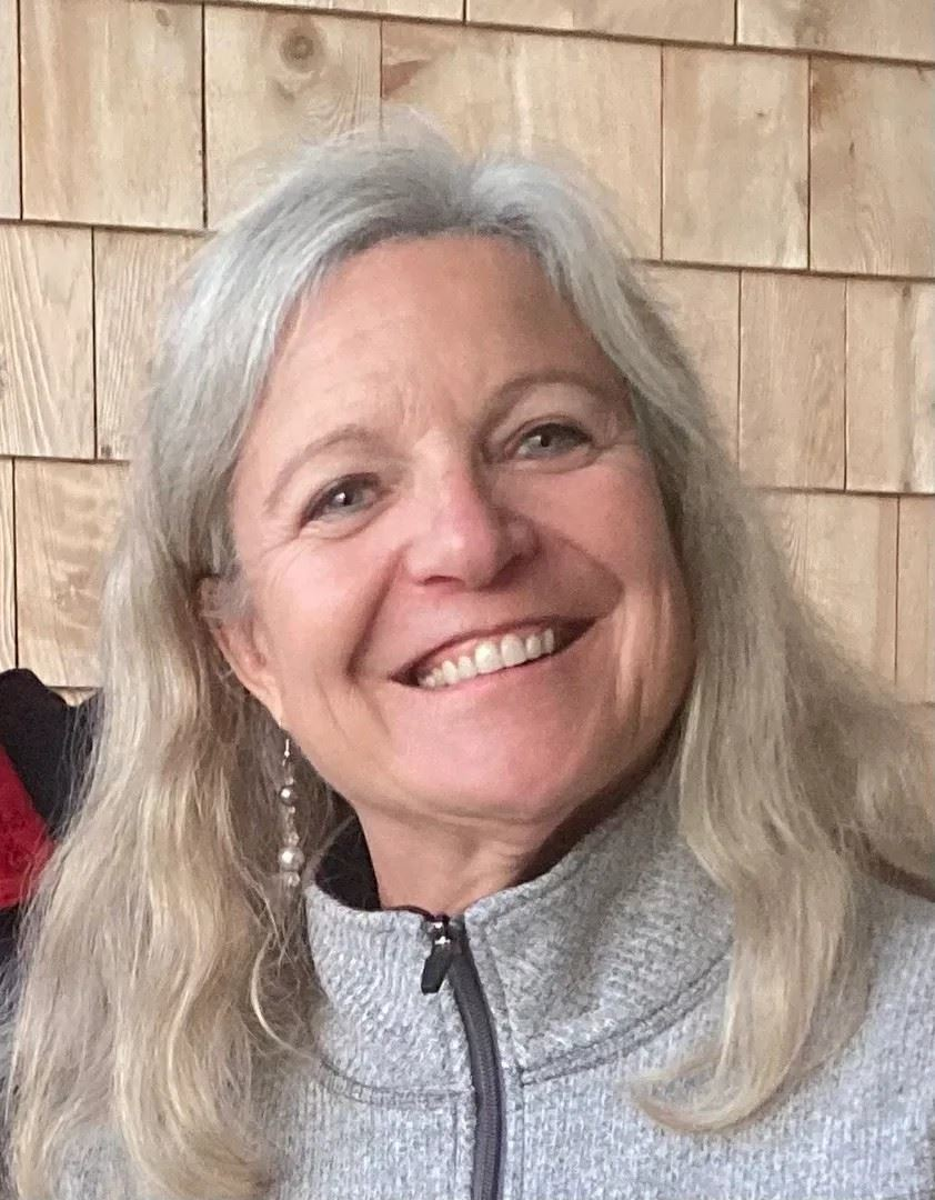 A woman with gray hair is smiling in front of a wooden wall.