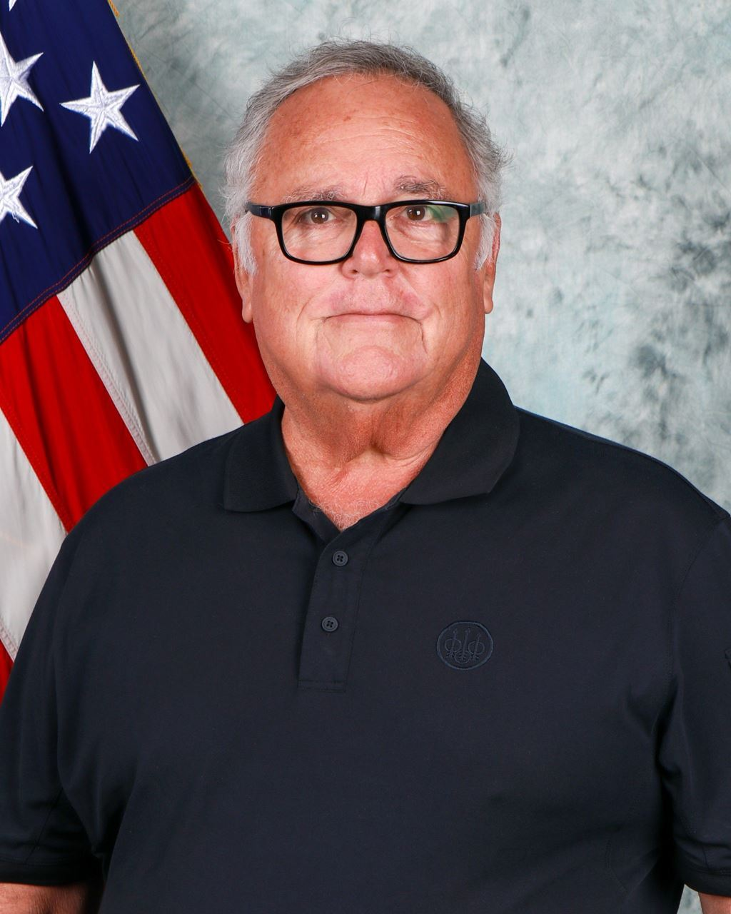 A man wearing glasses and a black shirt is standing in front of an american flag.