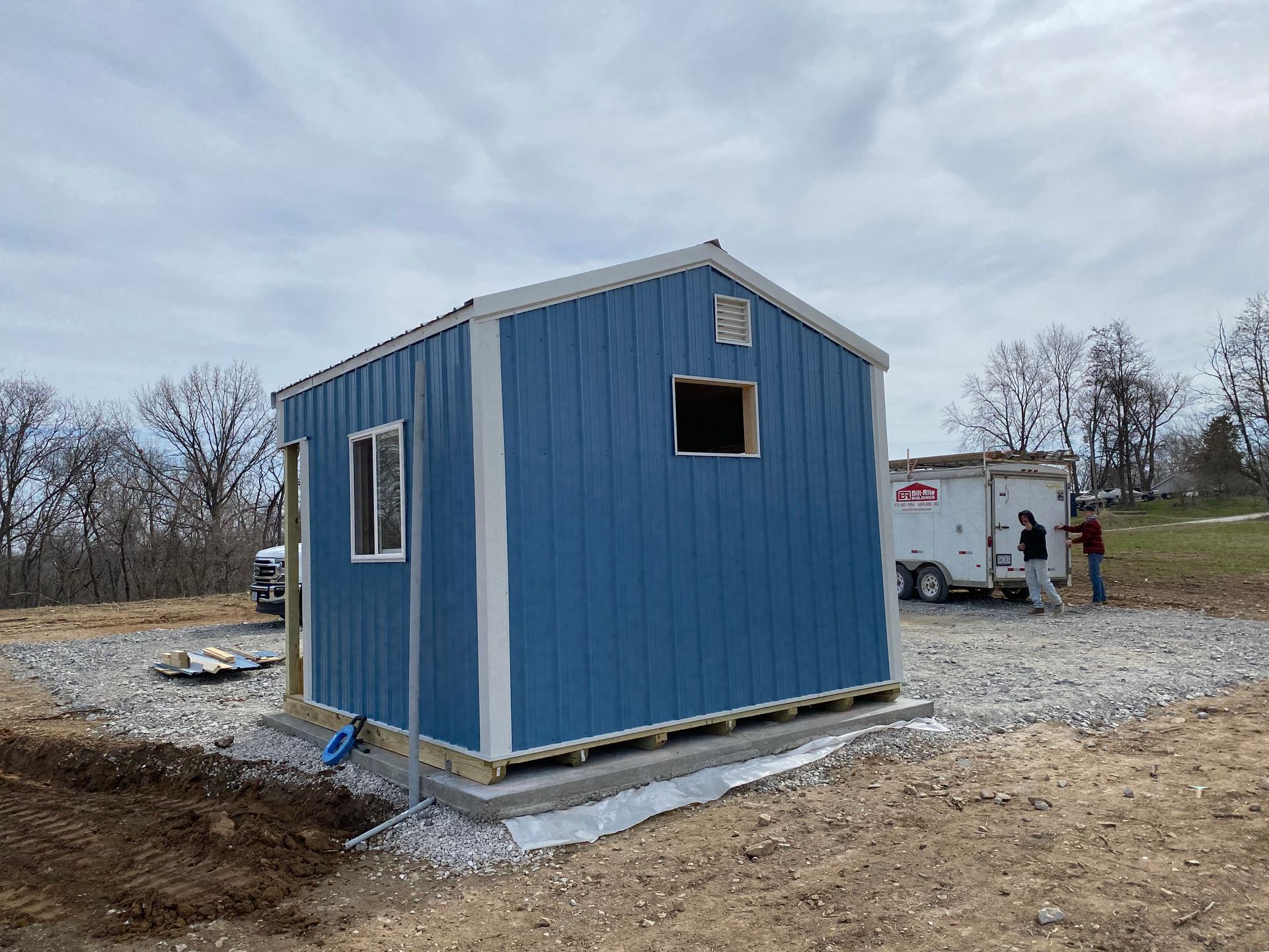 A small blue shed under construction, with a person nearby and a trailer.