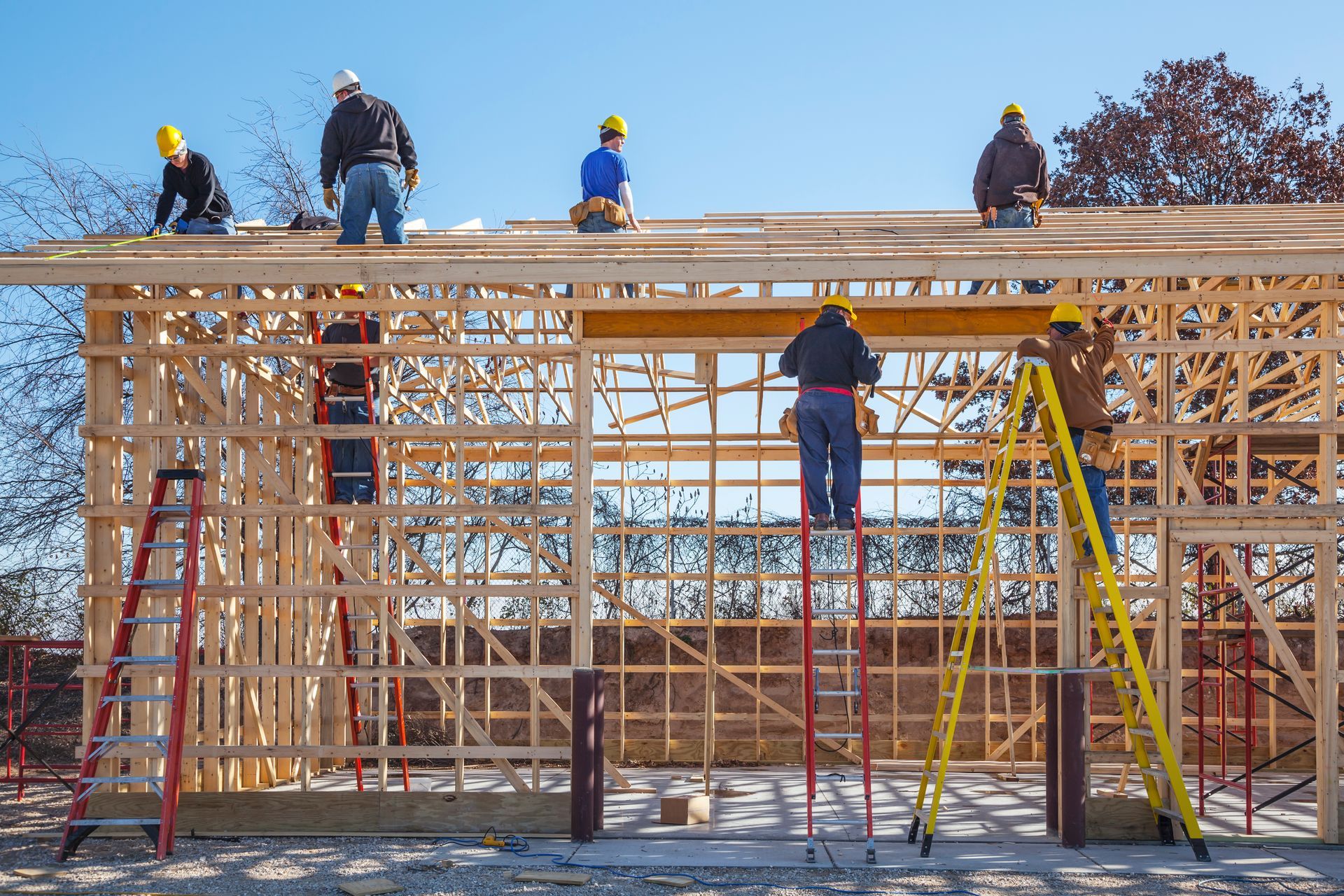 Construction workers build wooden frame using ladders and helmets, with trees and blue sky behind