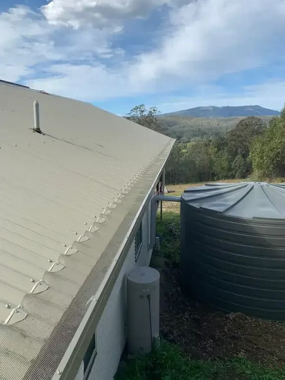 A light-colored house with a metal roof, gutter guard, and water tanks; mountain in the background.— Gutter Guard In Lake Cathie, NSW