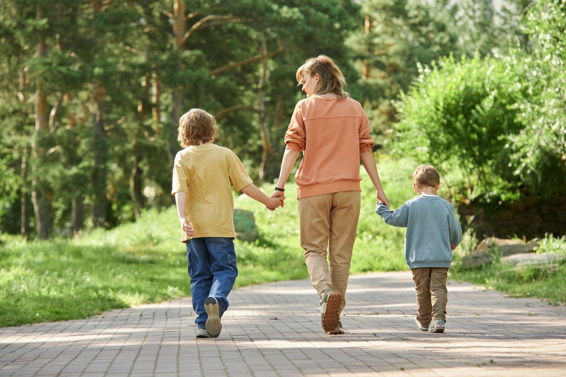 Three children holding hands and walking down a sunlit park path