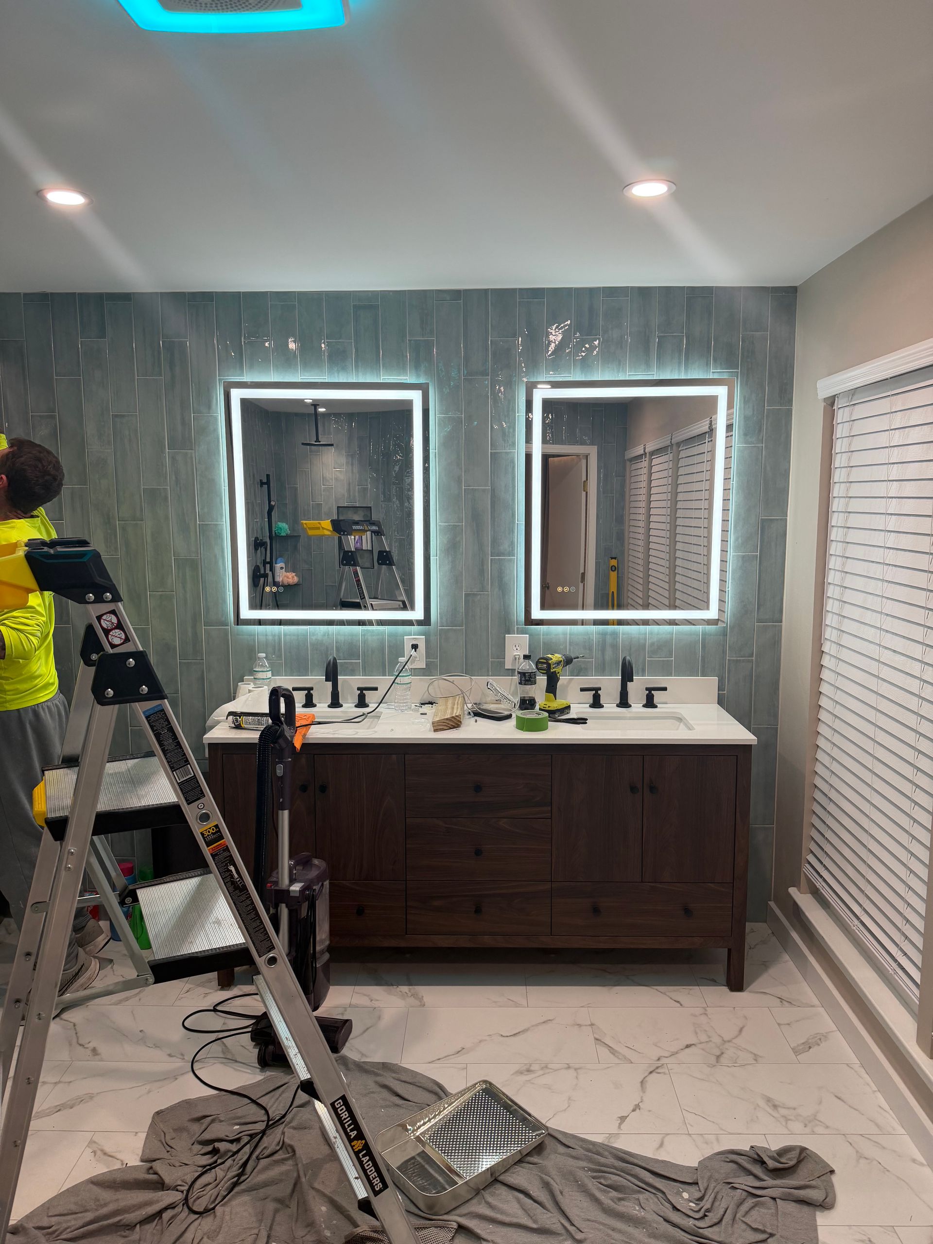 Bathroom with a double vanity, mirrors with LED lights, and a worker on a ladder.