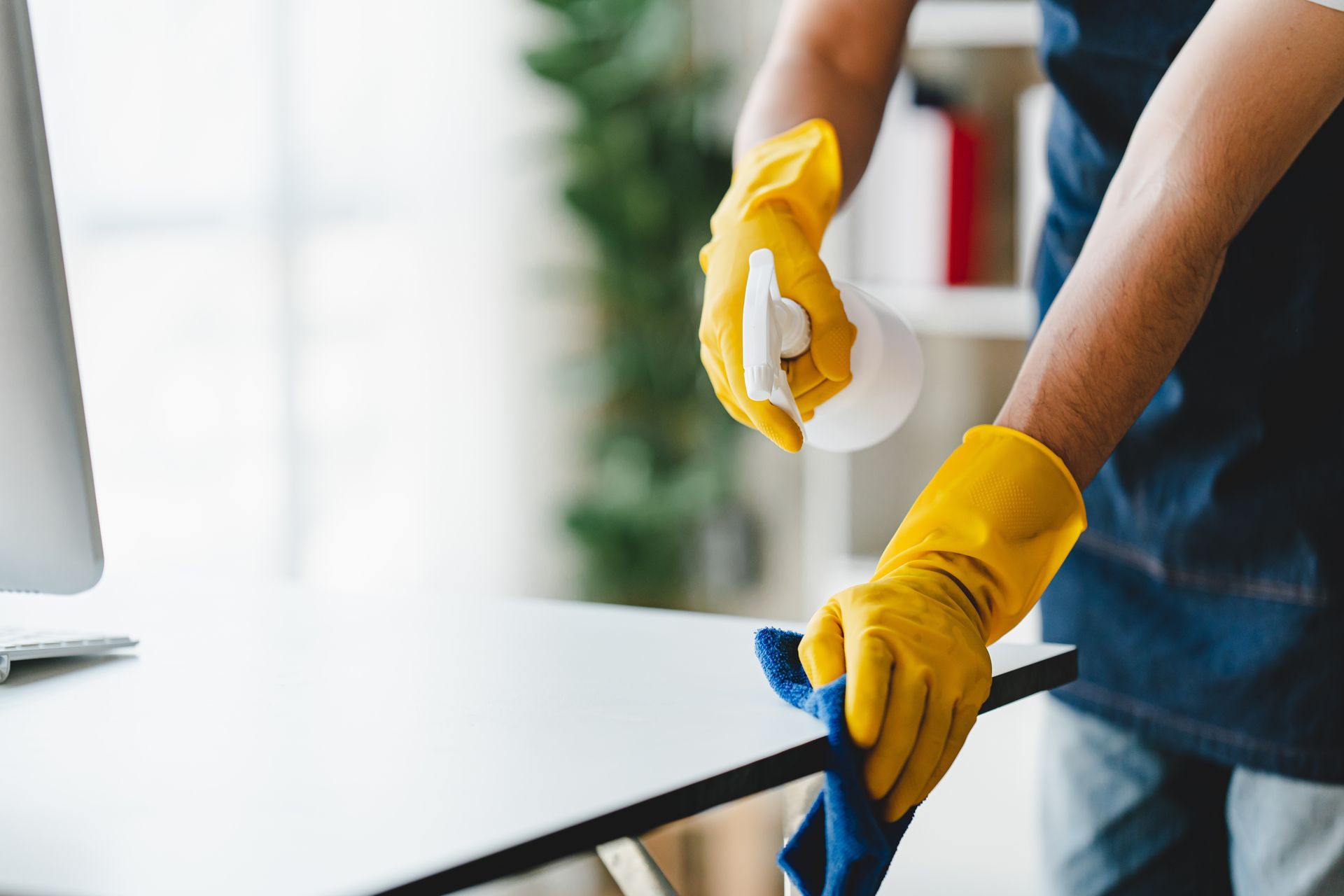 A person wearing yellow gloves is cleaning a desk in front of a computer